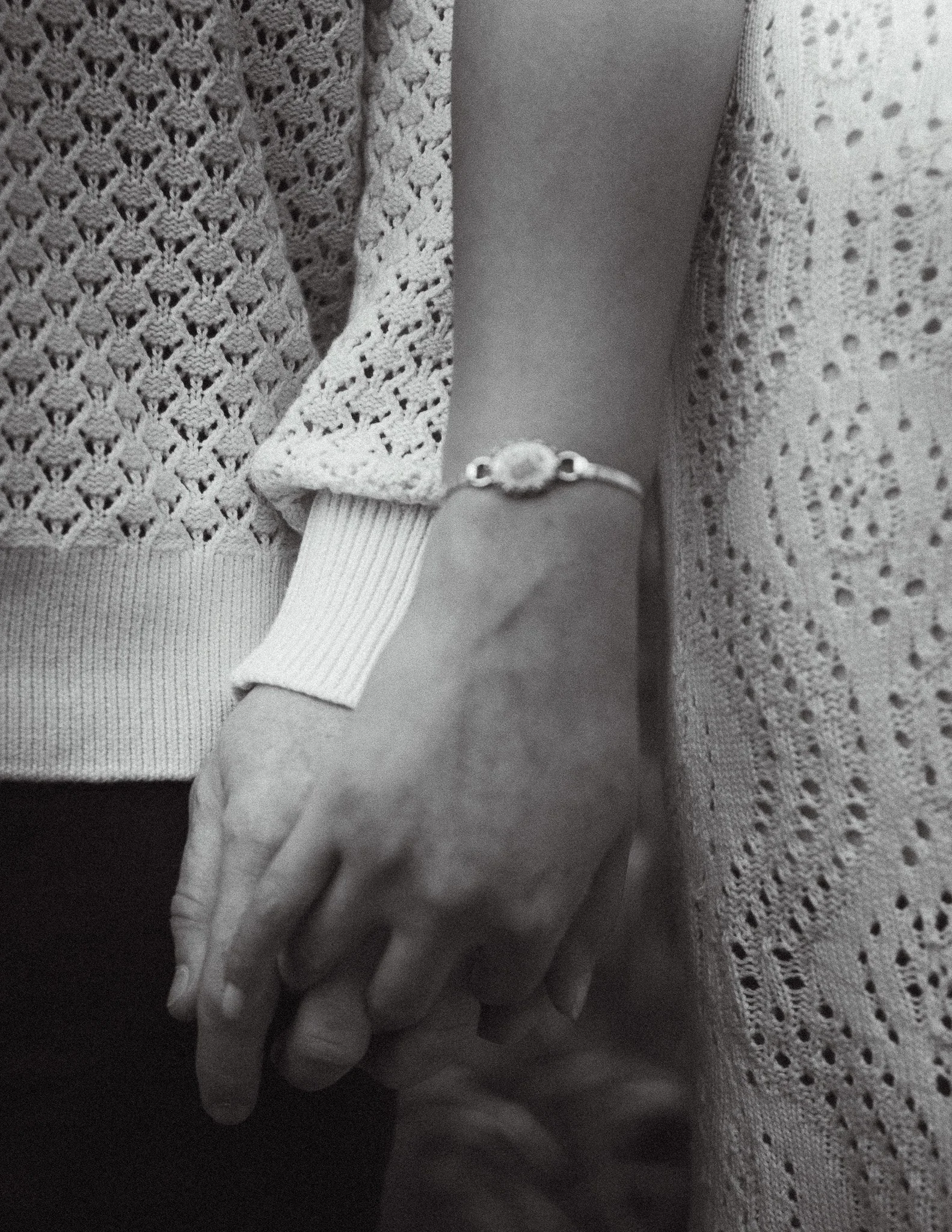A person's hand wearing a ring with a round stone, holding another hand. The background has a knitted or crocheted fabric with lace patterns, and there is fabric with holes on the right side of the image.
