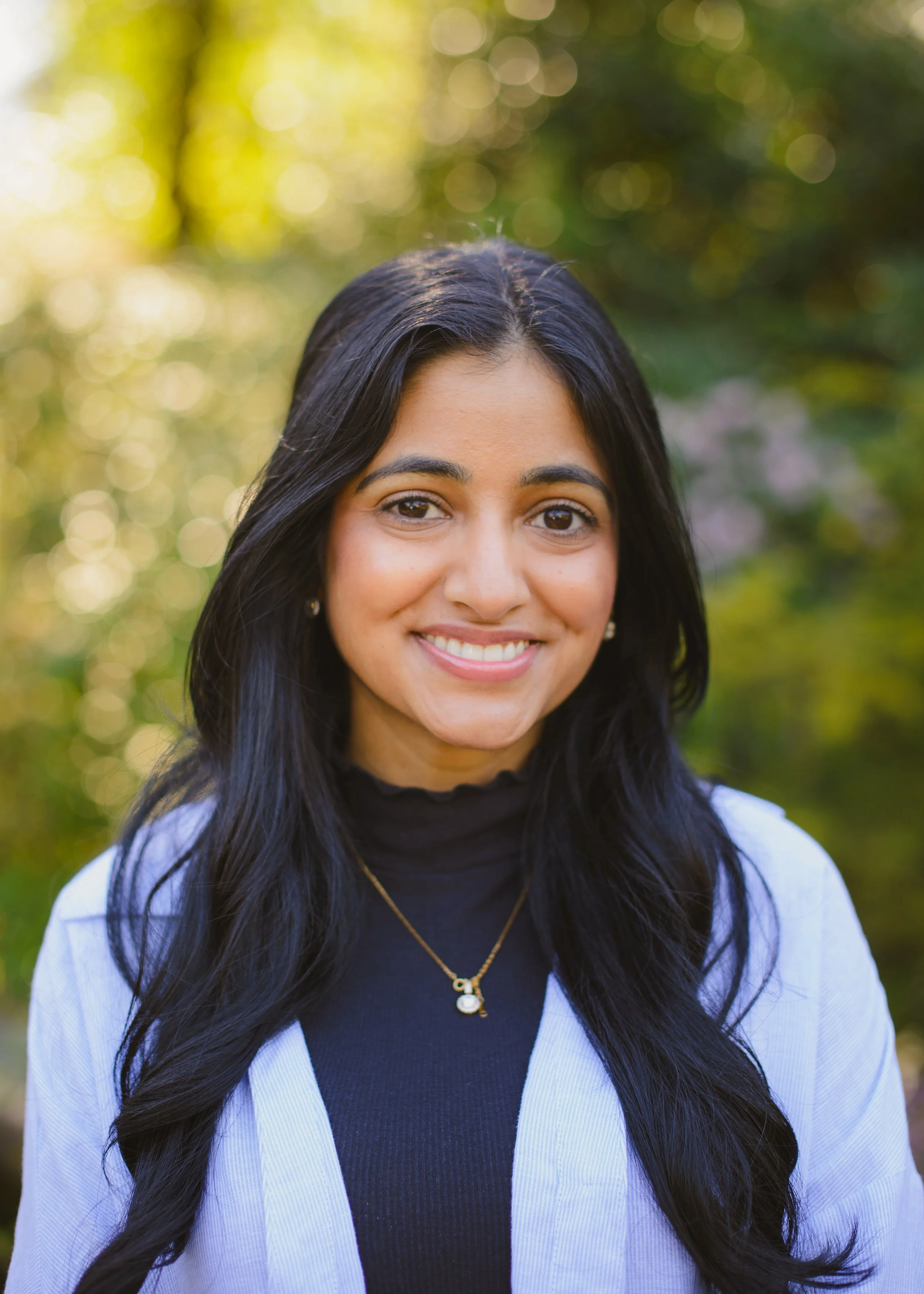 Close-up of a smiling woman with long dark hair wearing a black turtleneck, light-colored blazer, and a gold necklace, standing outdoors with blurred green and yellow foliage in the background. in bellevue Washington