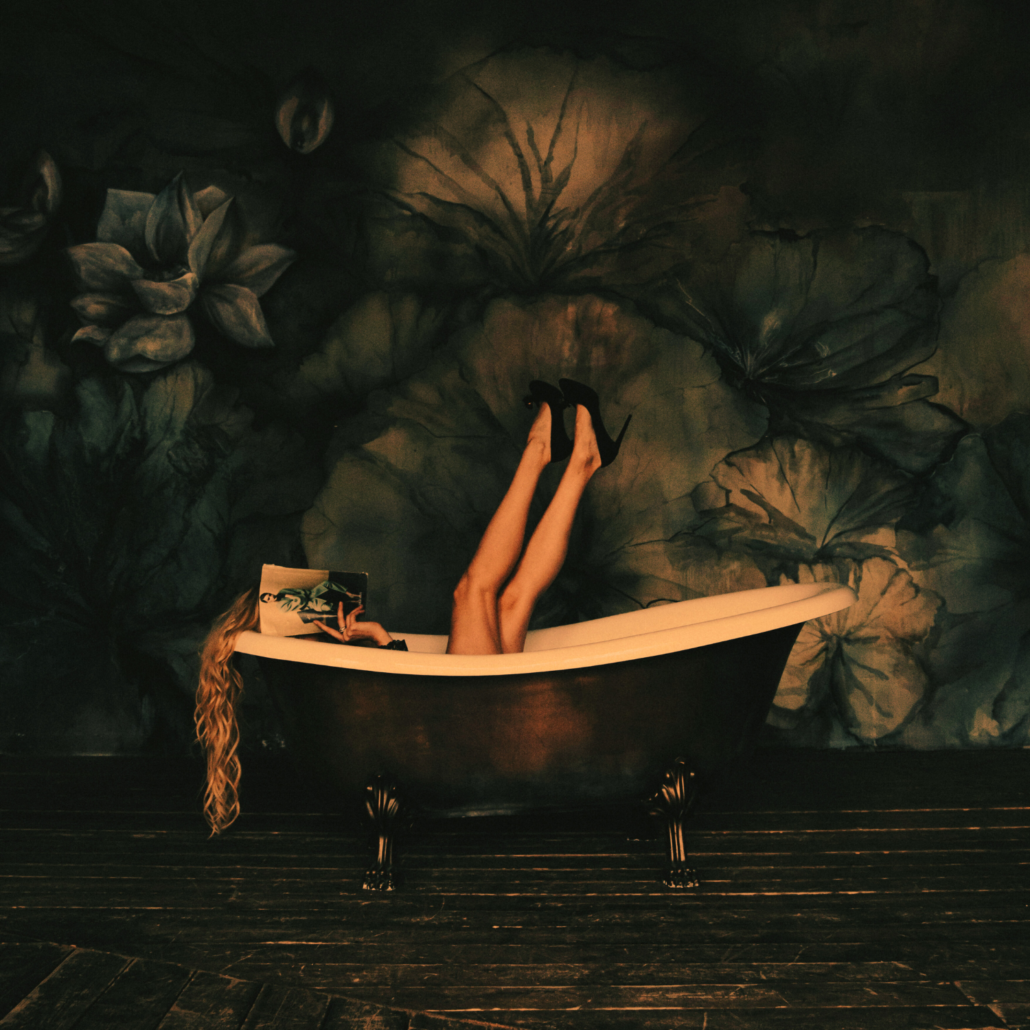 Person lying in a vintage clawfoot bathtub reading a book with legs up, set against a dark floral wall mural.