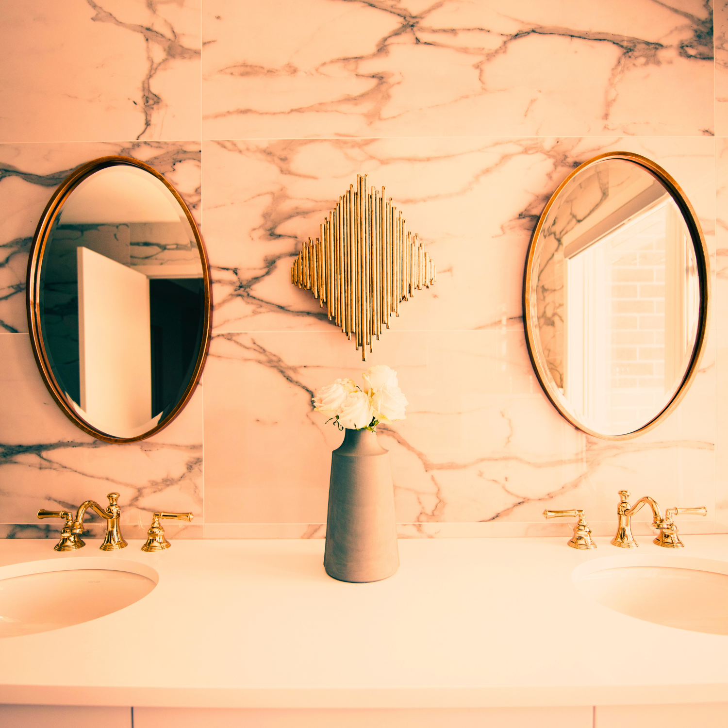 Bathroom vanity with dual oval mirrors, gold fixtures, a marble wall, a ceramic vase with white roses, and a gold decorative wall art piece between the mirrors.
