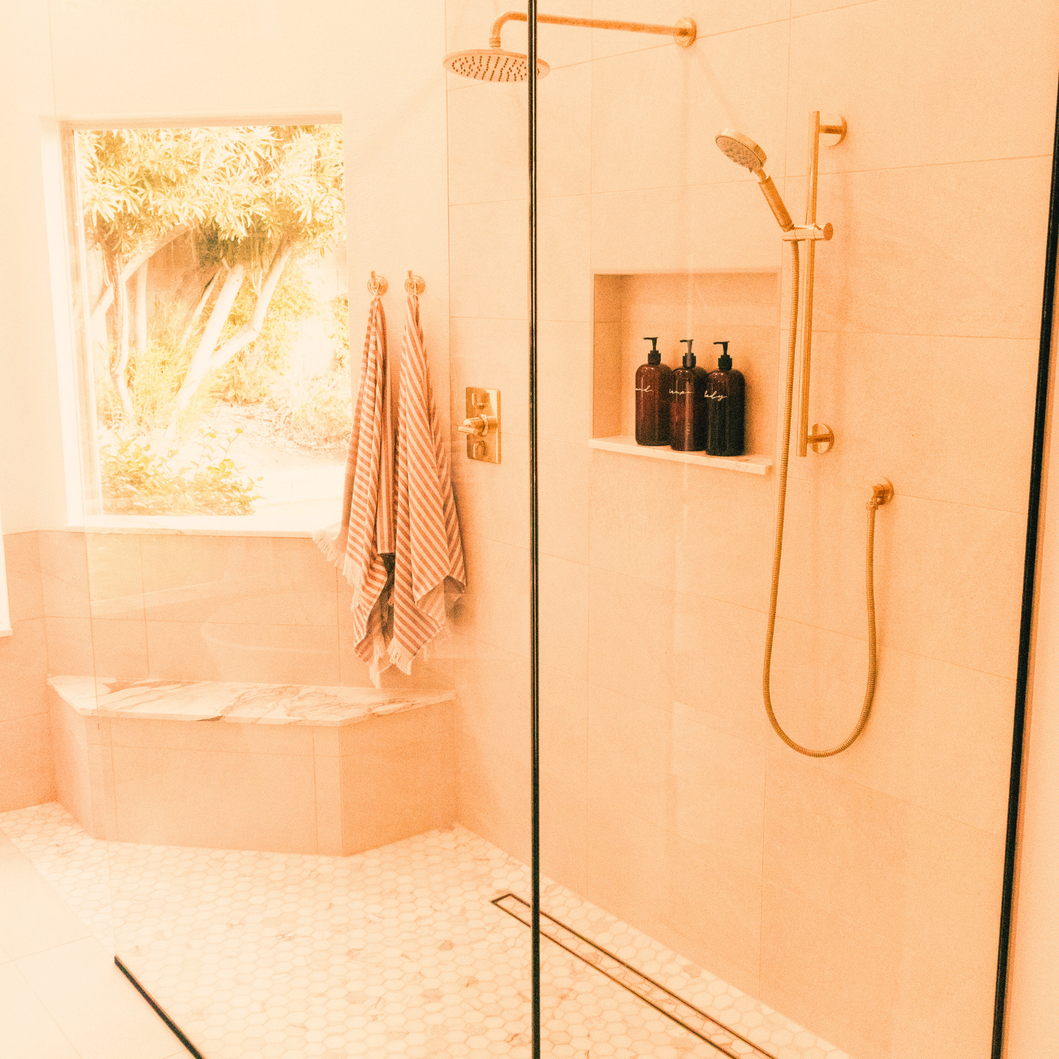 Bathroom shower with beige tiles, rainfall and handheld showerheads, a built-in shelf with three dark soap dispensers, a tiled bench near a window with a view of trees outside, and two striped towels hanging on hooks.
