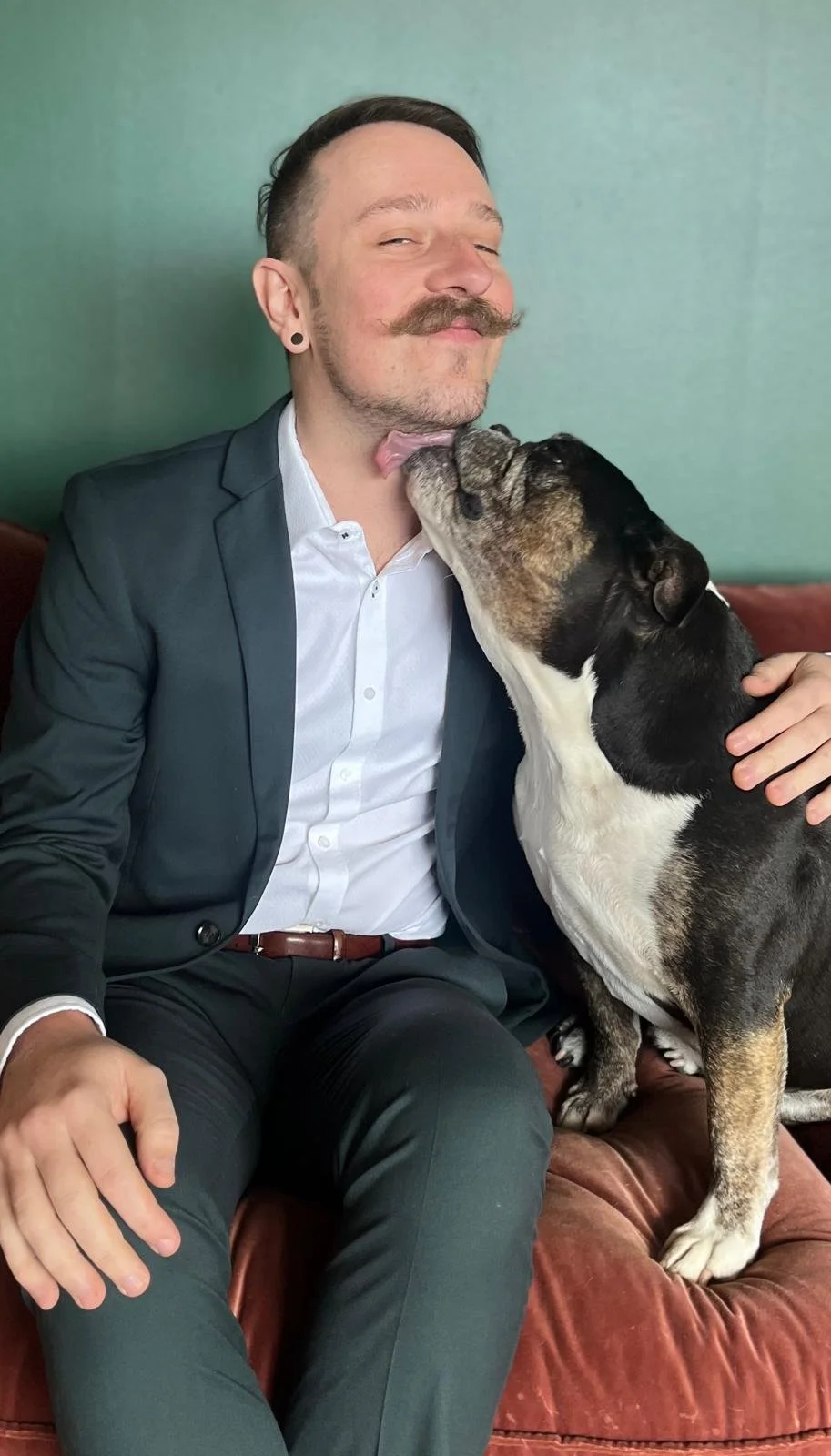 Man in a suit with mustache sitting on a couch smiling as a bulldog licks his chin.