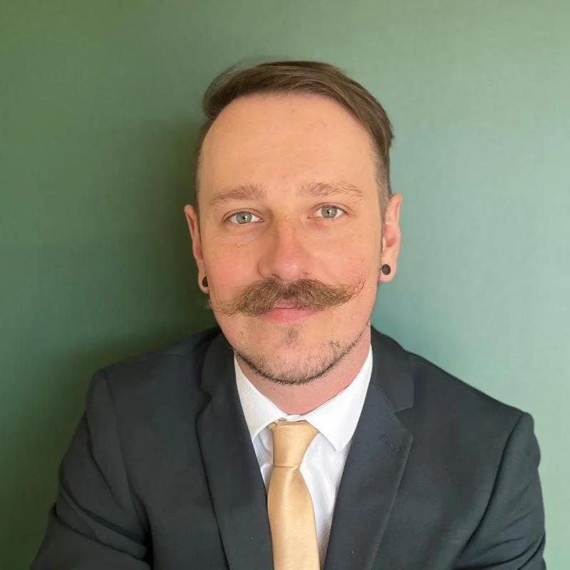 Man with a mustache wearing a black suit and beige tie against a green background.