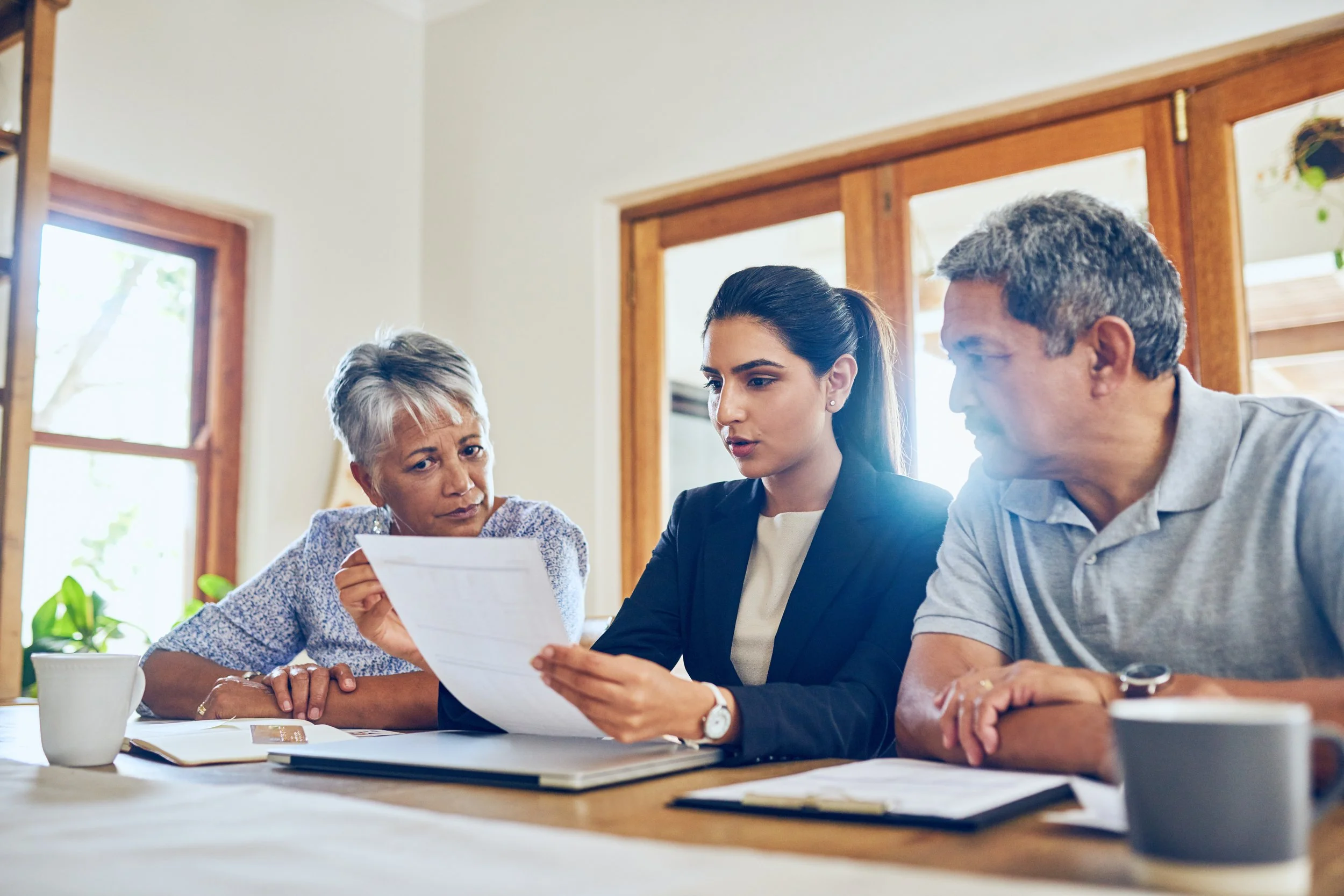 A professional woman discussing paperwork with an older couple at a table, with a laptop and documents present.