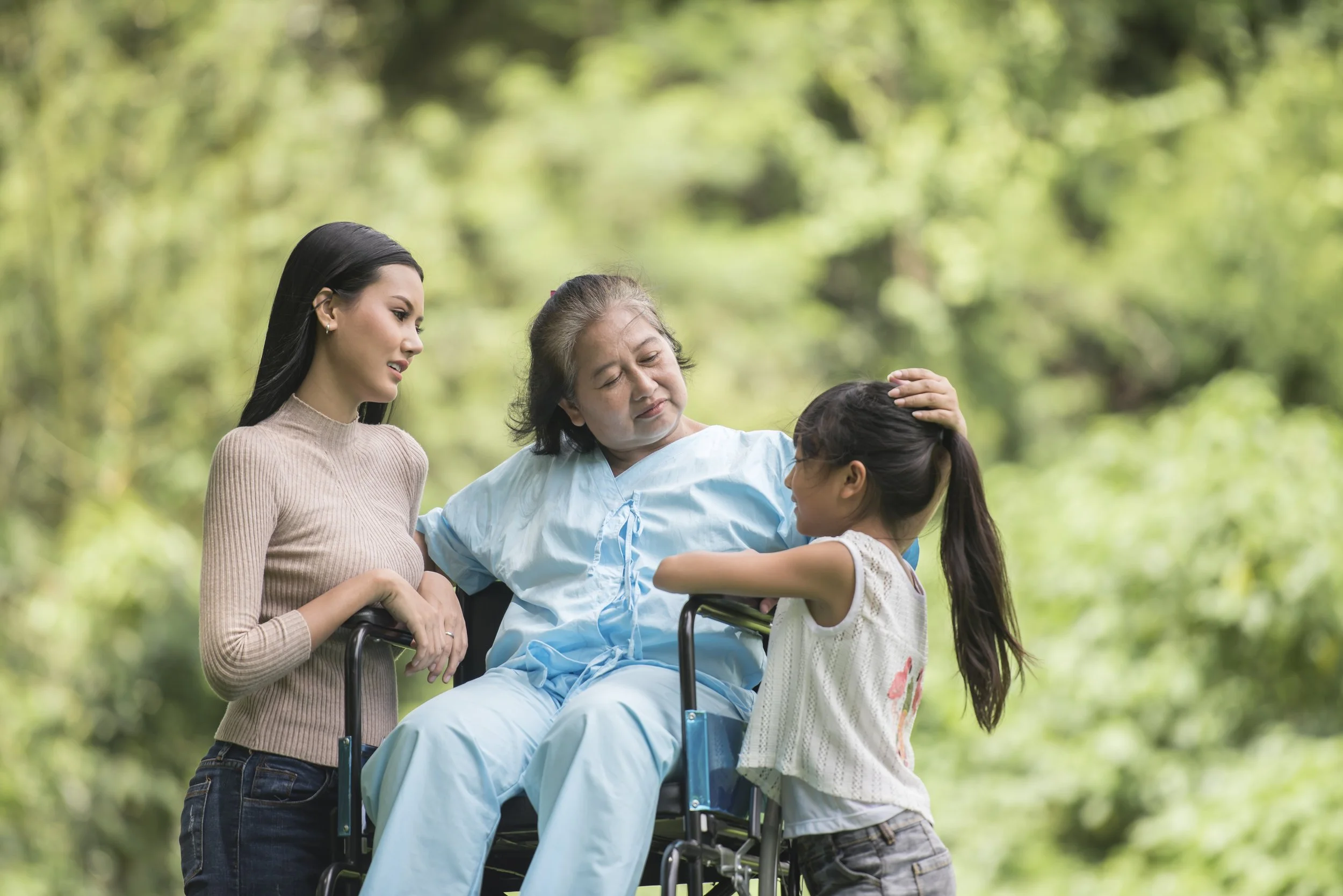 Three people outdoors, with a middle-aged person in a wheelchair, a younger woman standing beside them, and a child smiling and interacting with the seated person. The setting is a green park.
