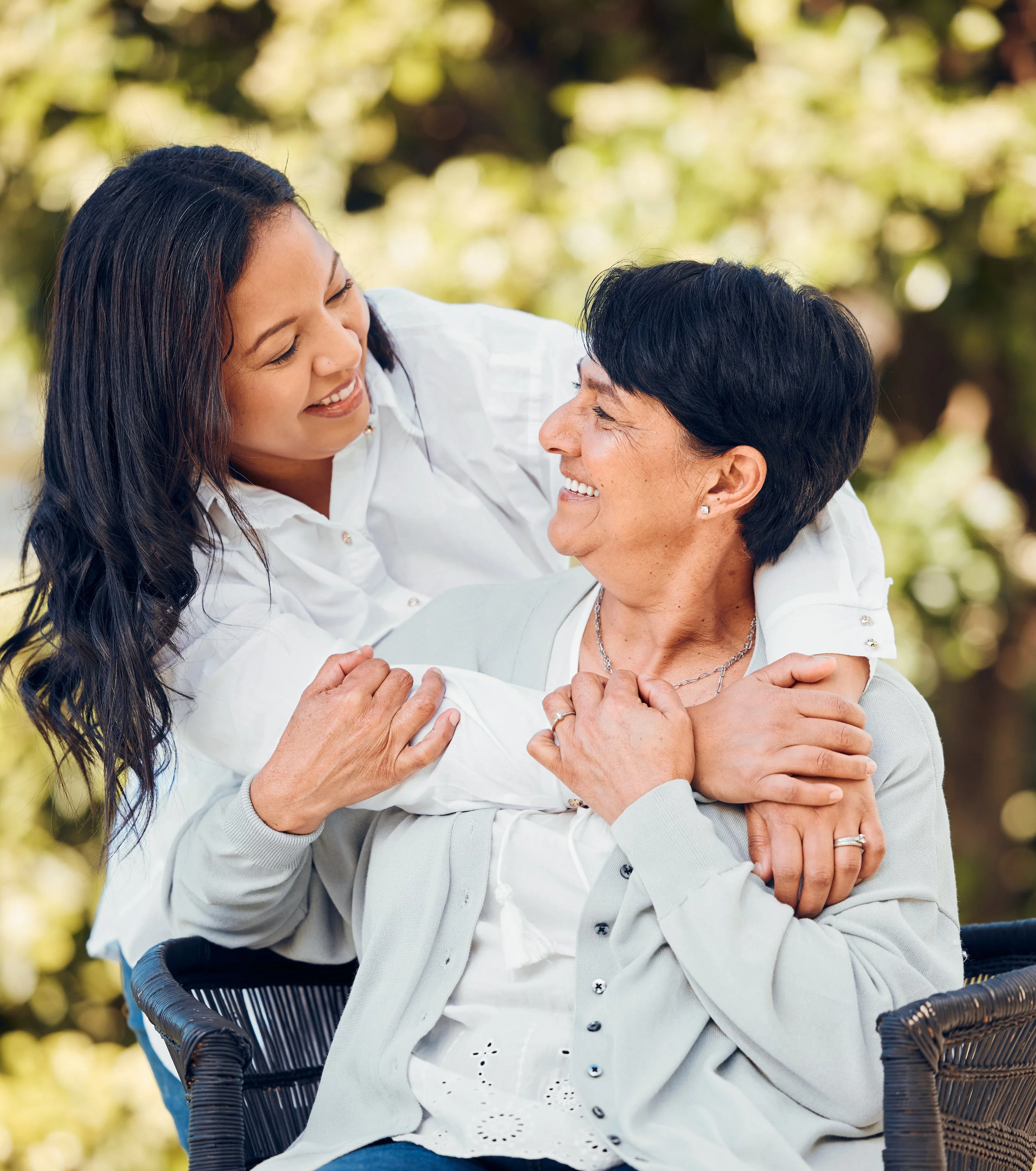 Two women smiling and embracing outdoors, with one sitting and the other standing behind her.