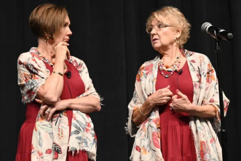 Two elderly women talking to each other on a stage, both wearing red dresses and floral shawls, with a microphone nearby.