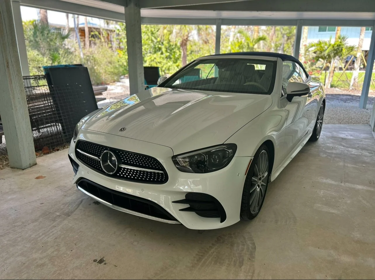 White Mercedes-Benz convertible car parked in a covered area with plants and trees in the background.