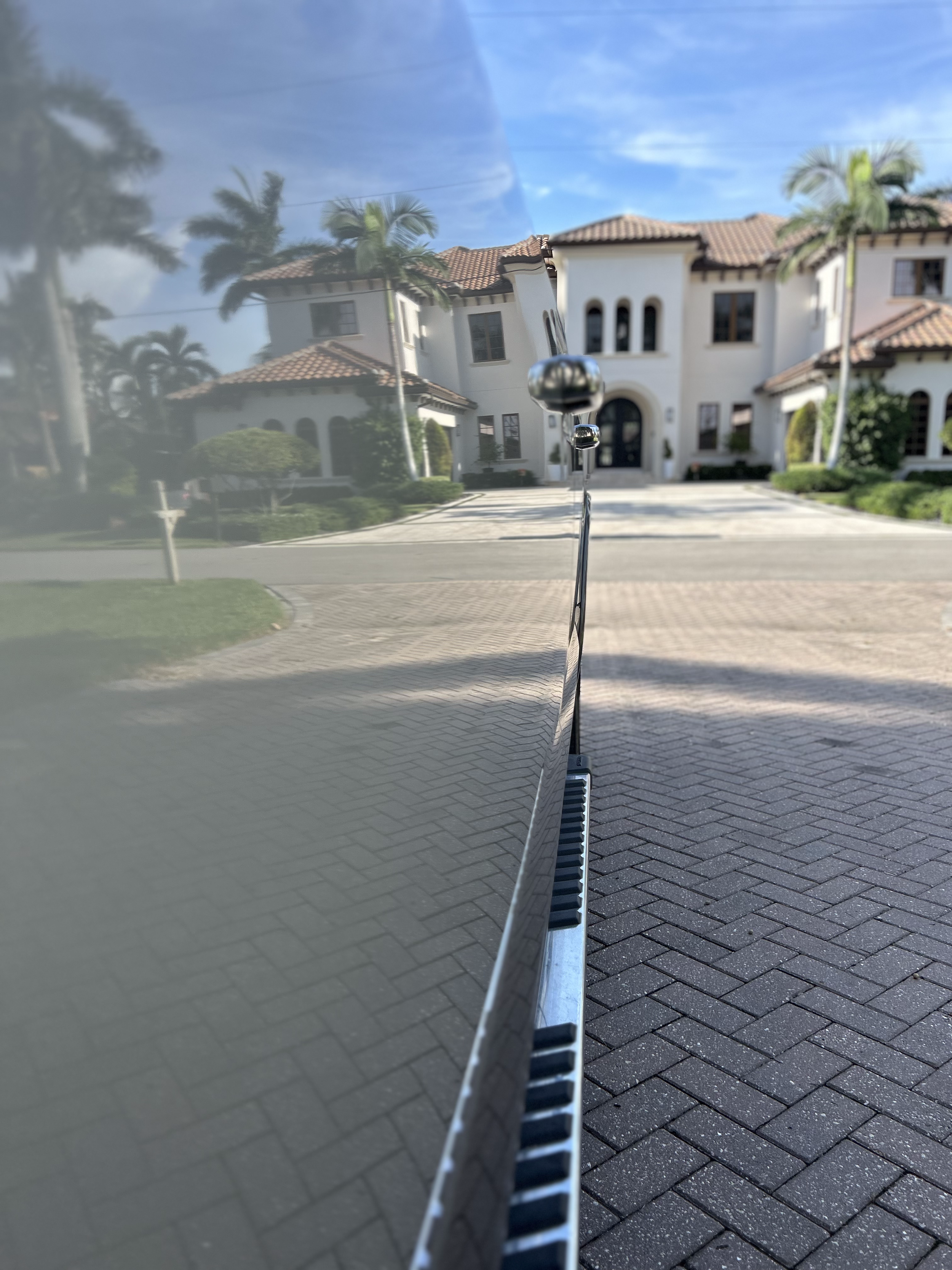 A reflection of a house with a red tiled roof, white walls, palm trees, and a clear blue sky visible on the side mirror of a black car parked on a paved driveway.