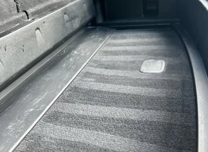 The image shows the footwell of a vehicle, with a textured gray floor mat, a metal footrest, and a part of the vehicle's side panel.