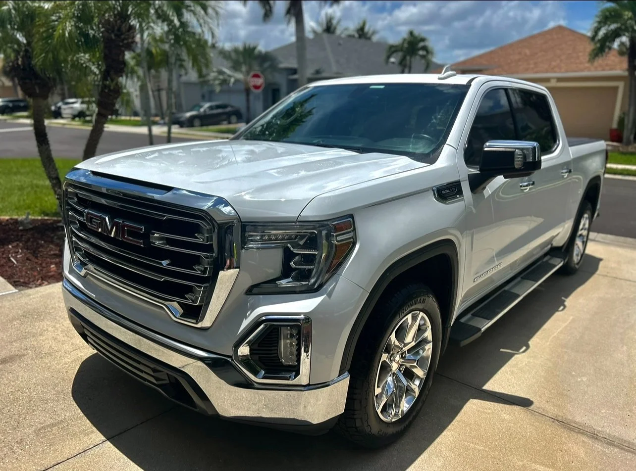A silver GMC Sierra pickup truck parked on a driveway in a suburban neighborhood with houses, palm trees, and a stop sign in the background.
