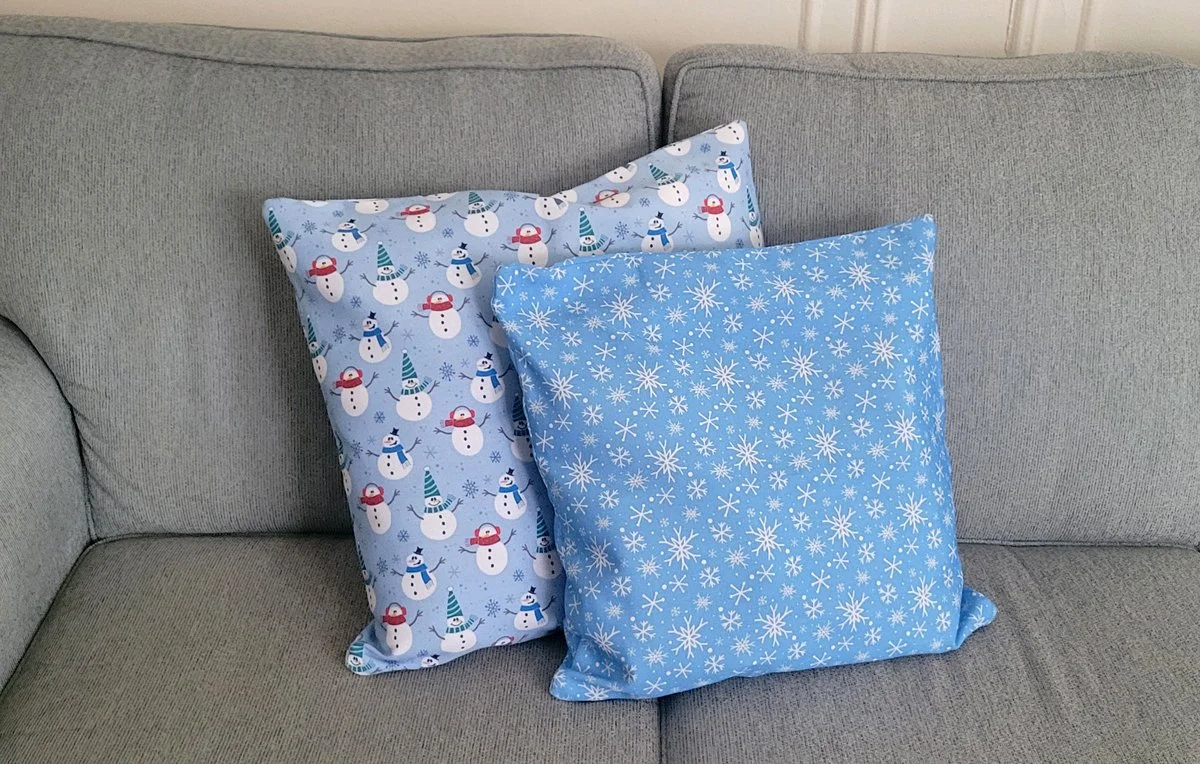 Two decorative holiday pillows on a couch featuring patterns with snowmen and snowflakes