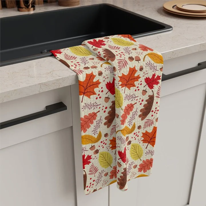 A tea towel hanging over a kitchen sink with a fall pattern featuring colorful autumn leaves, twigs, ferns, and acorns