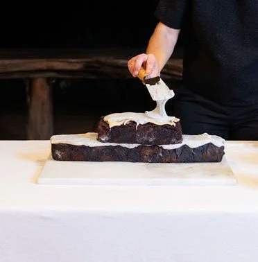 Trowel applying grey mascarpone between chocolate brick layers, live performance, edible installation construction.