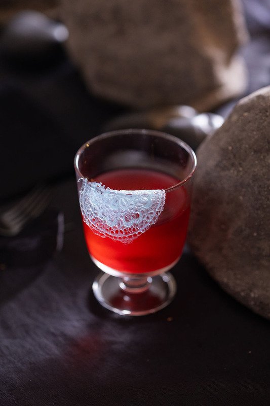 Edible lace in butterfly pea blue folded at the rim of a red elixir glass.