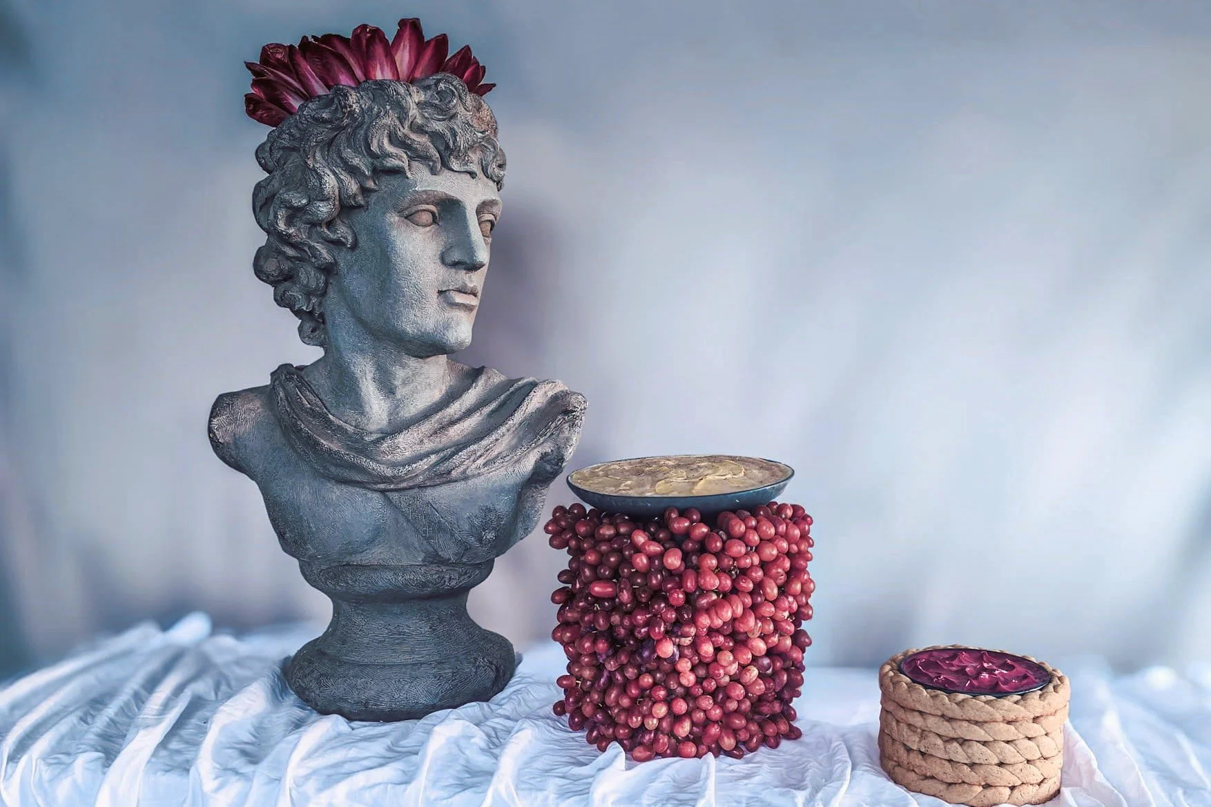 VECETO: Food still life, grey bust, tower of grapes and braided bread wreaths on white linen. Pink in all three.
