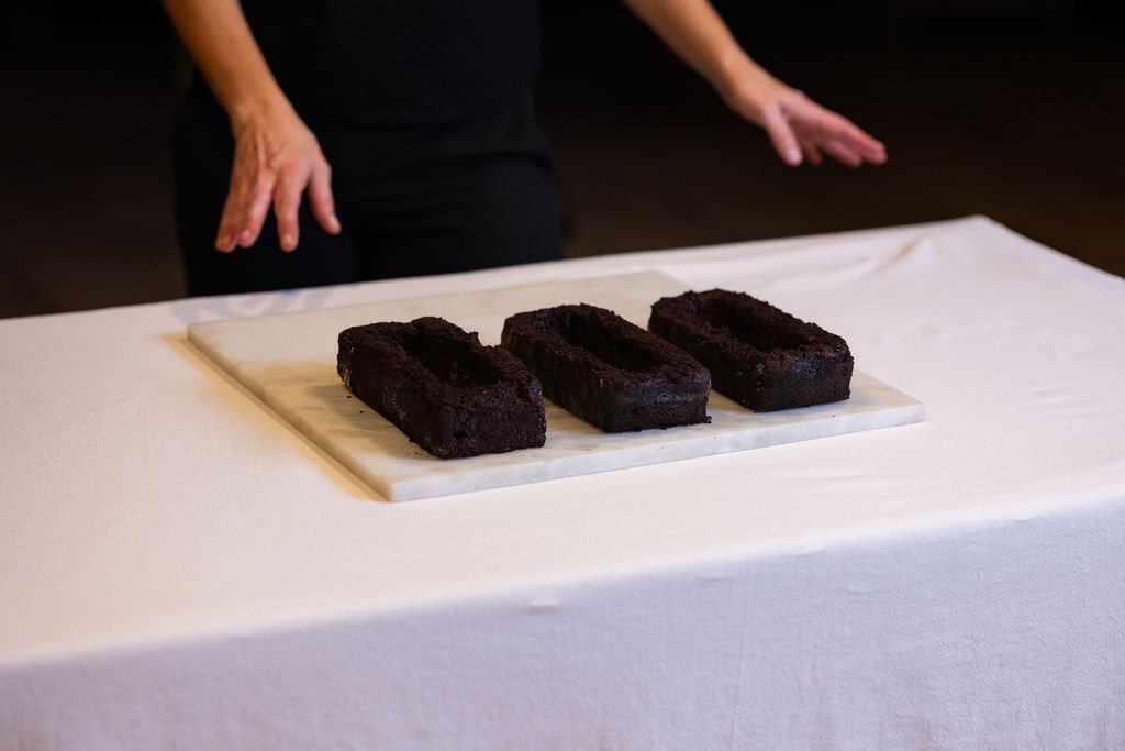 Three hollow chocolate cakes on marble surface, hands gesturing toward composition, live performance documentation, spatial gastronomy