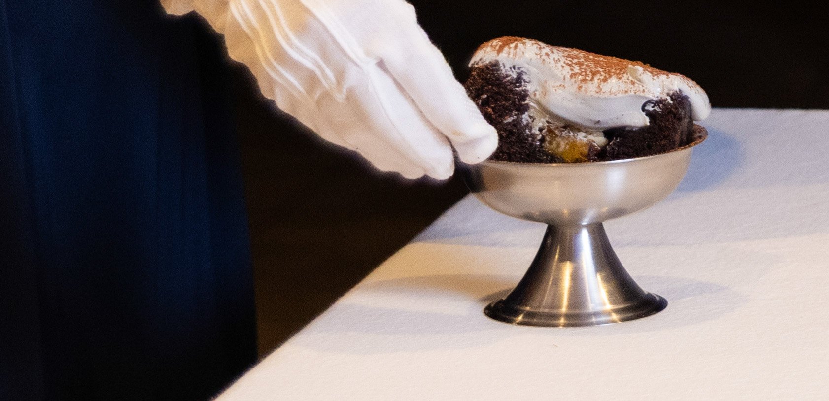 White gloved hand cradling silver dessert bowl from behind, ceremonial service, sensory dining experience, black ground.