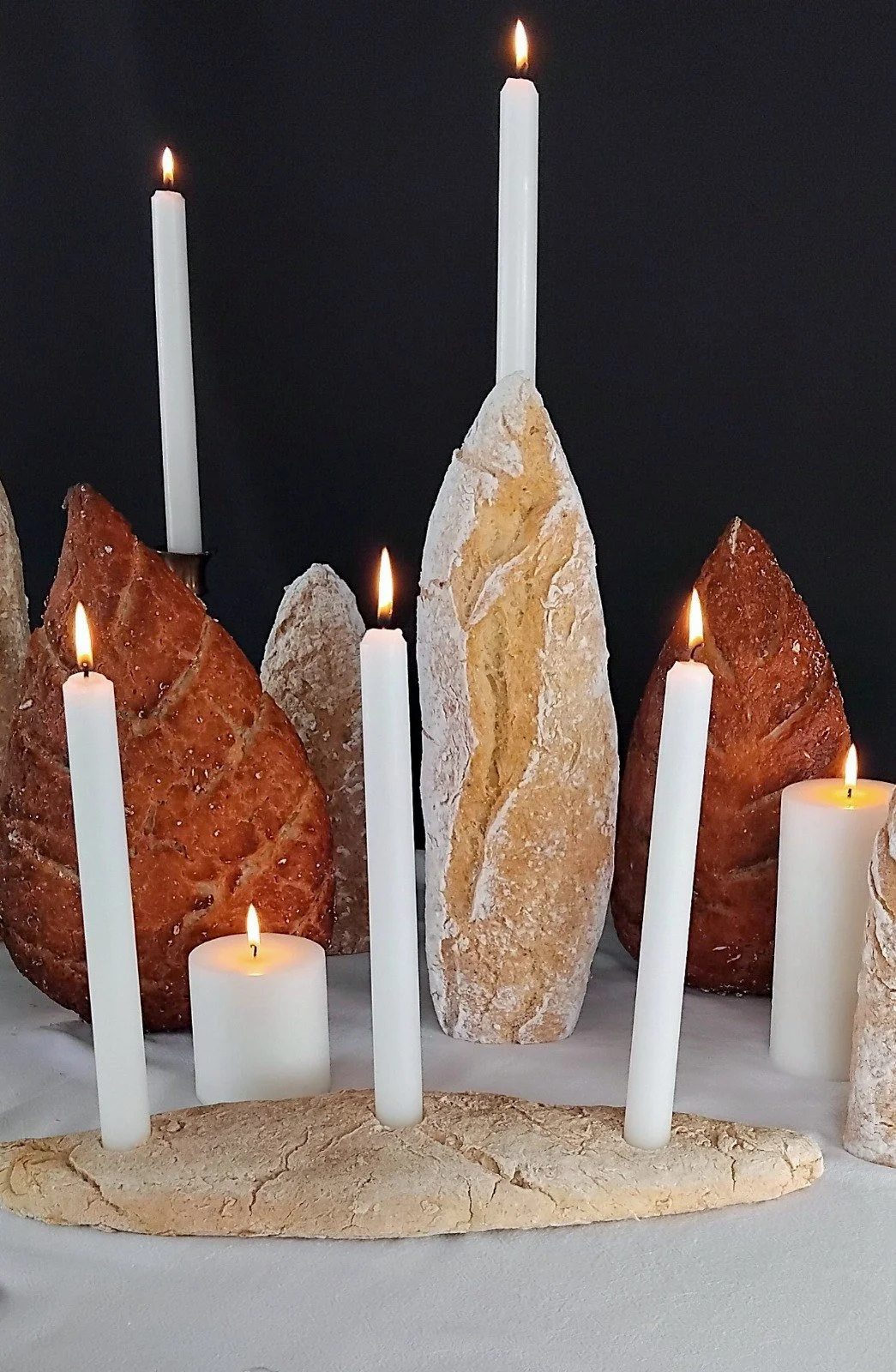 VECETO: Ephemeral sculpture, standing loaves among lit white candles on cream linen, dark ground.