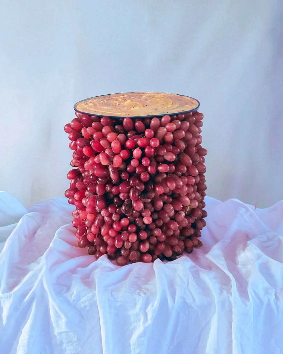 VECETO: Gastronomic still life, tower of pink grapes under a dark grey bowl of gratin, white linen below.