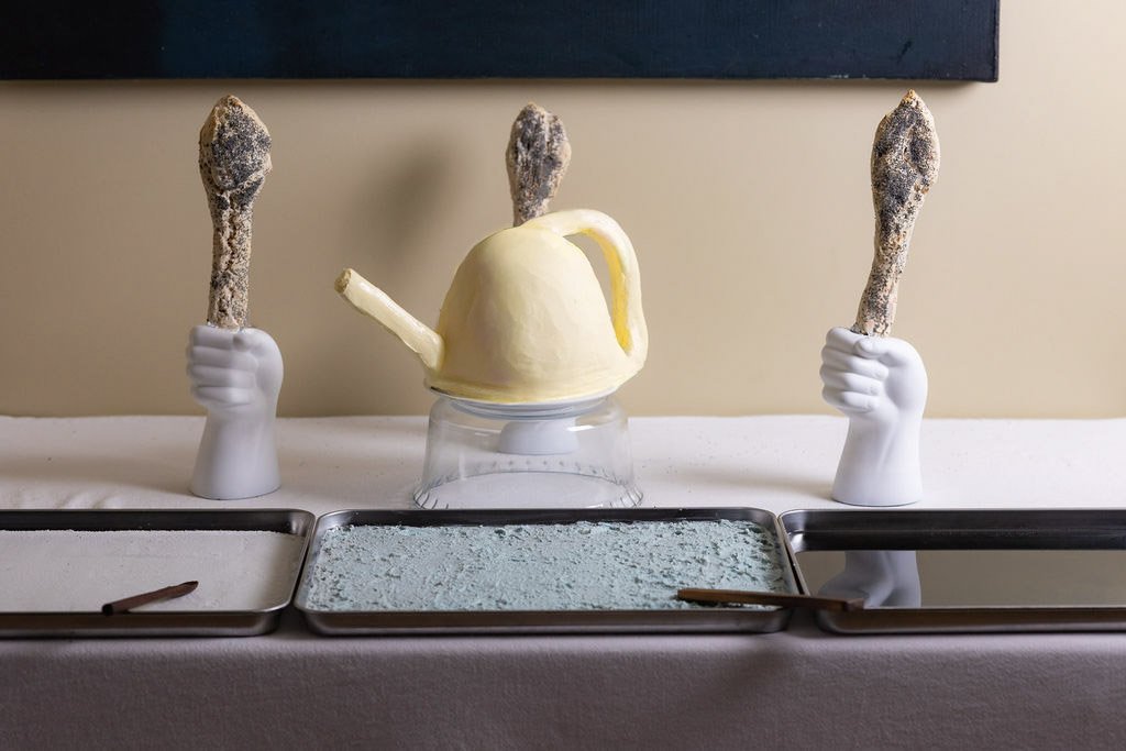 Ceremonial tableau: three blue trays, bread sceptres in ceramic hand vessels, butter watering can sculpture at centre, spatial gastronomy.