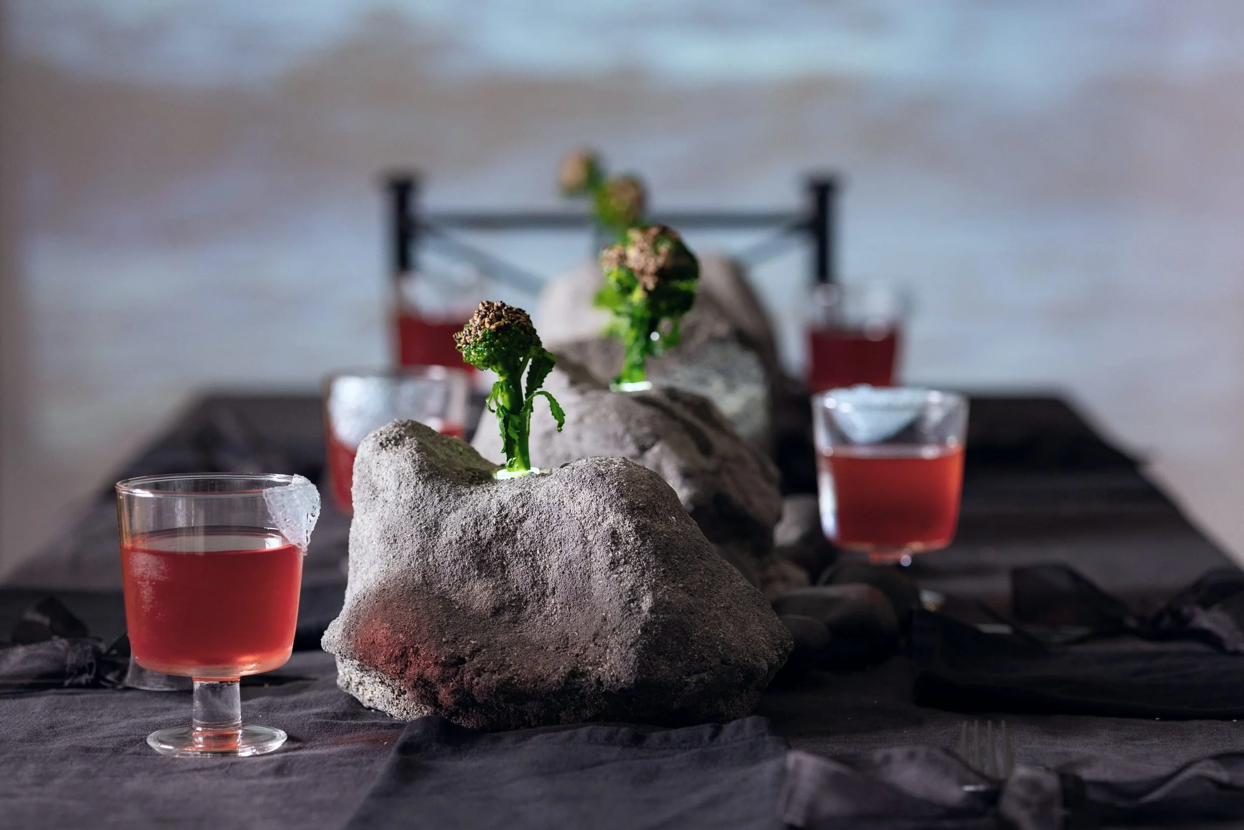 VECETO: Immersive installation art, concrete vessels and broccolini. Red elixir glasses before an ocean projection.