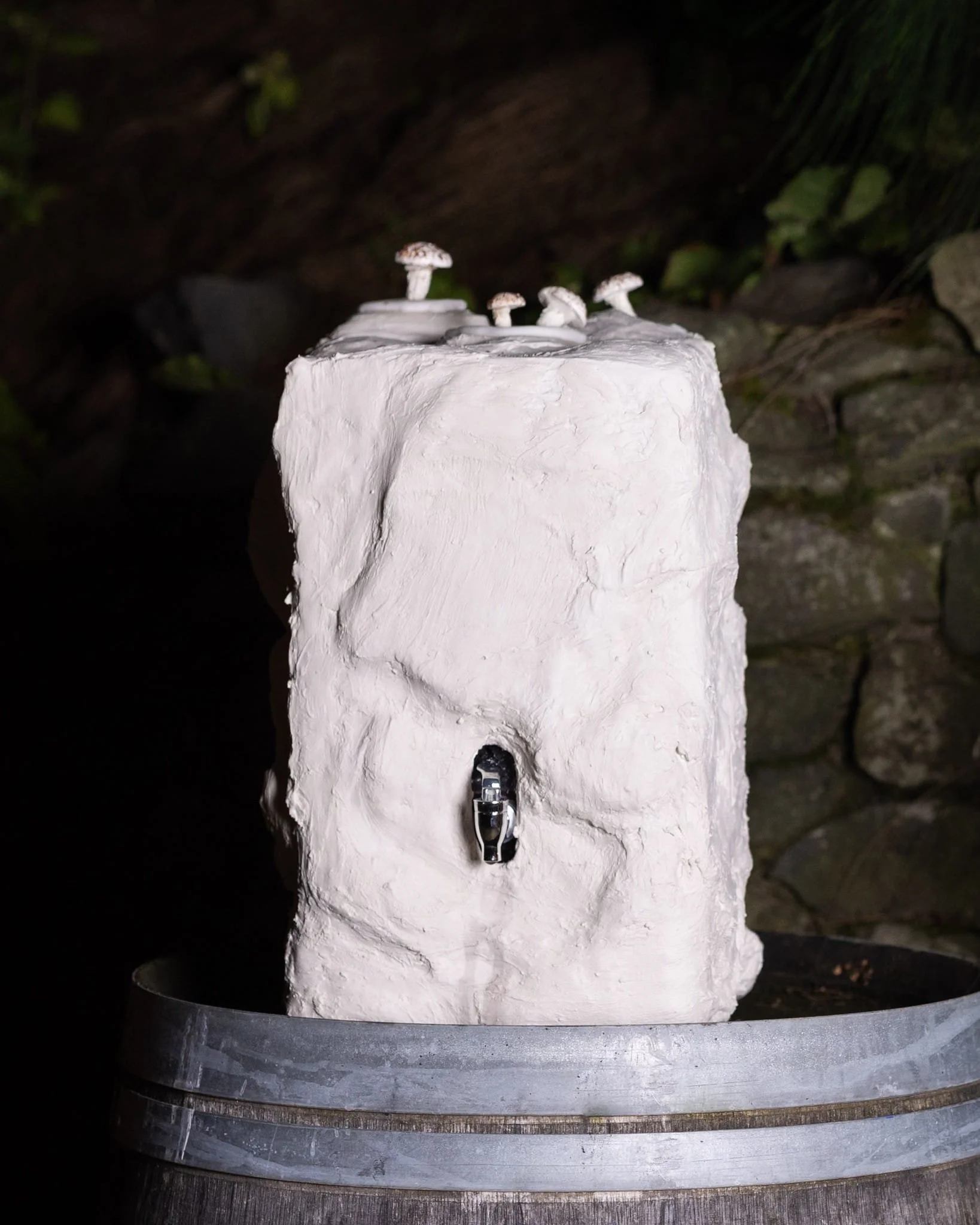 Food sculpture artists. Gypsum-clad functional vessel on wine barrel with confectionery mushrooms against stone wall.
