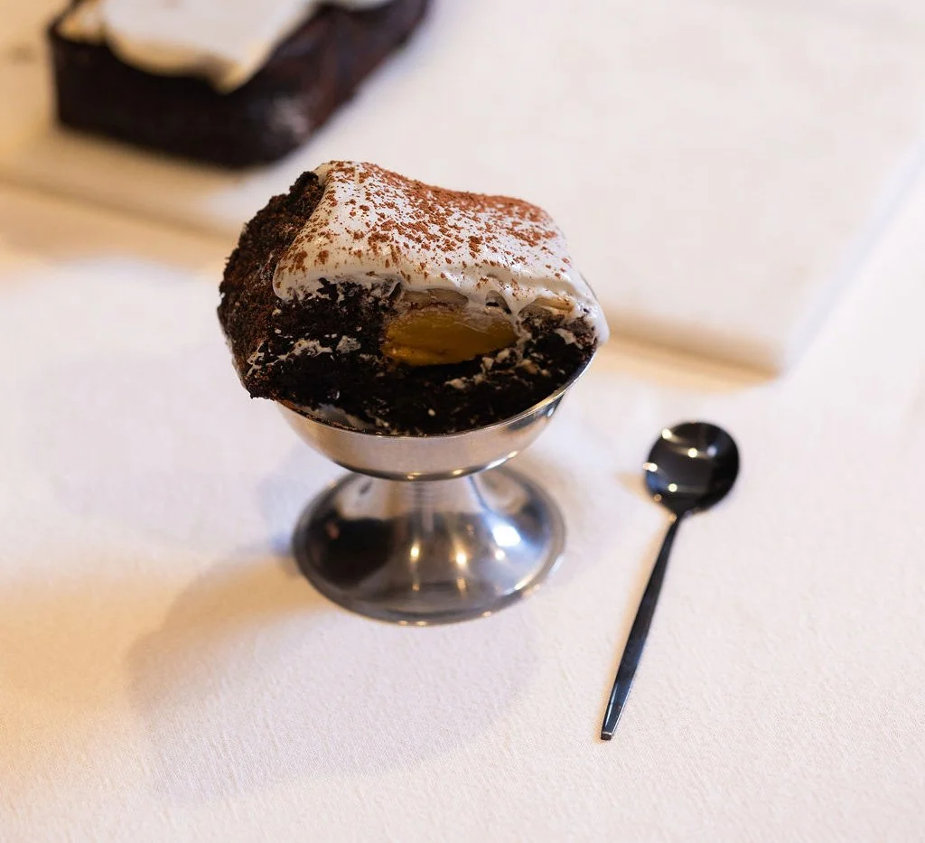 Silver dessert bowl with chocolate mascarpone layers, clinker wall visible behind, edible sculpture served, ephemeral installation.