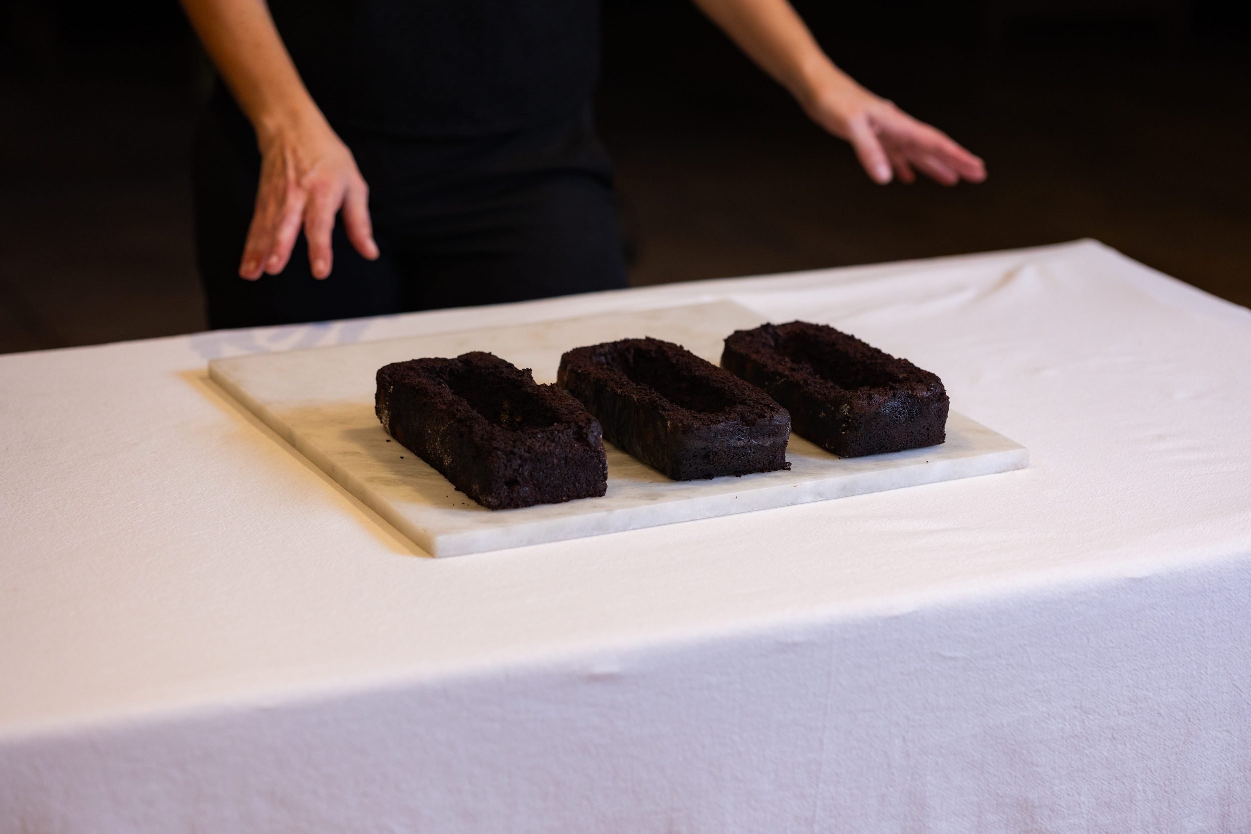 VECETO: Brutalist cake, hollow chocolate blocks on white marble. Dark sponge laid out before stacking, hands behind.