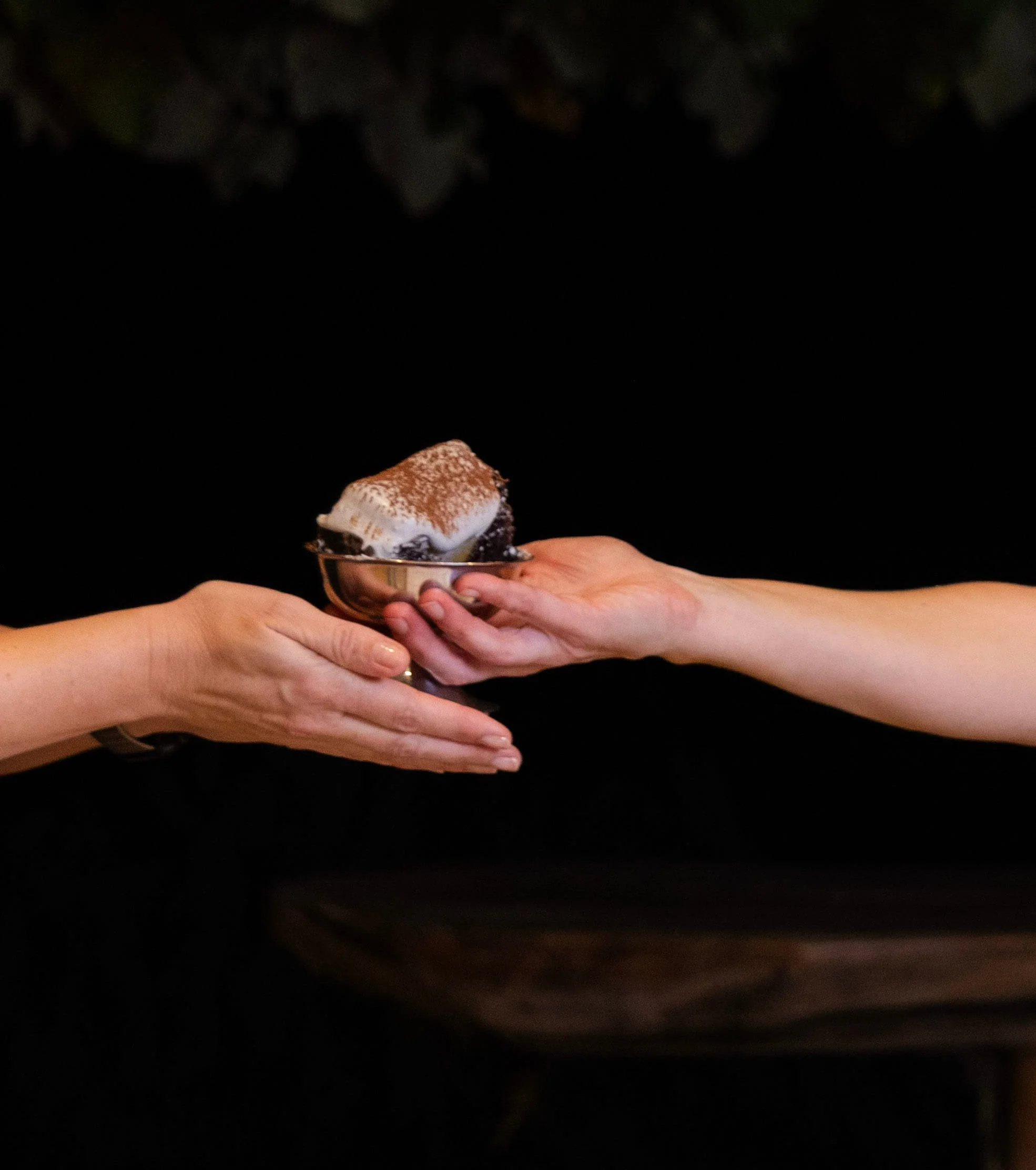 Two forearms in mid-air handover of silver bowl, mascarpone cacao surface, black background, performance culmination.