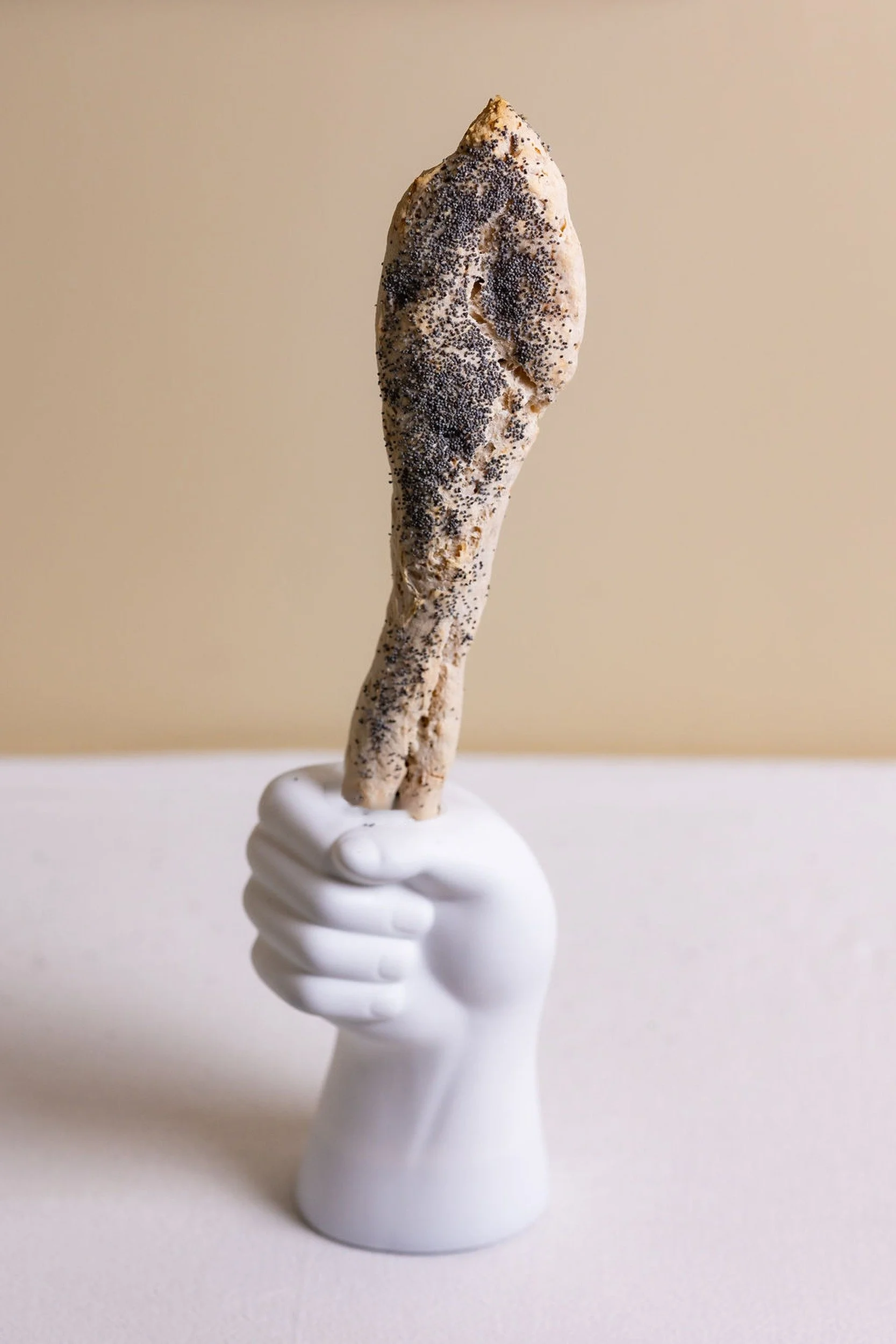 VECETO: Food performance art, bread sceptre with poppy seed held in a white ceramic fist against a warm sand wall.