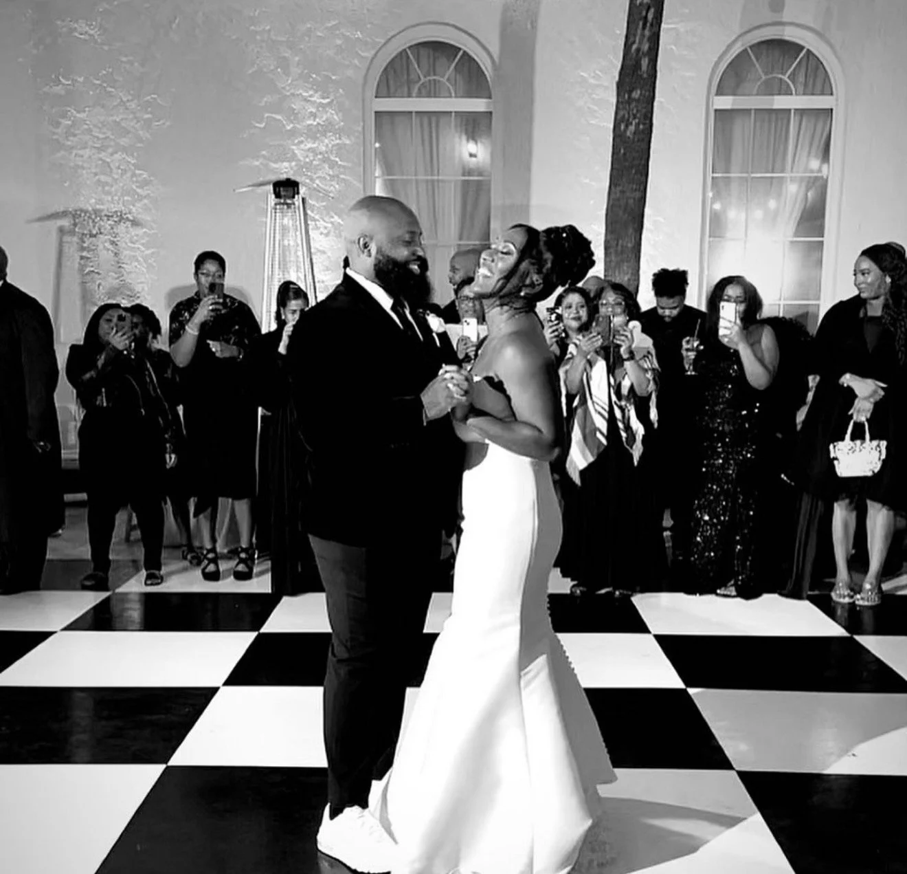 A black-and-white photo of a couple dancing at a wedding reception with guests in the background. The couple is smiling and looking at each other, with the woman wearing a strapless gown and the man dressed in a suit. Guests are taking photos and watching them with smiles, some holding drinks.