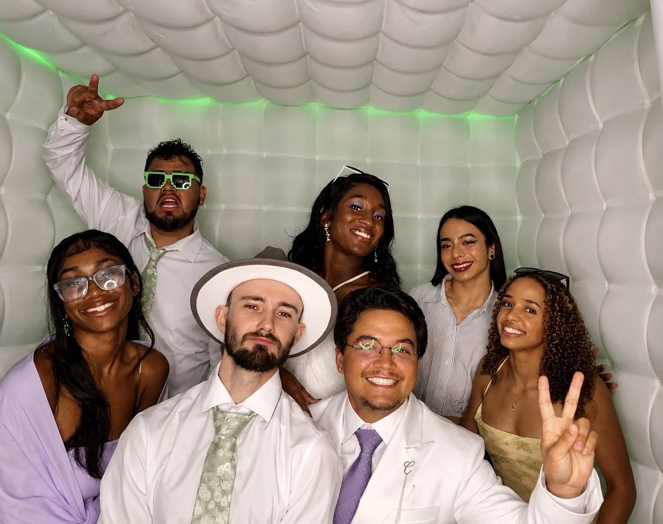 Group of friends celebrating in photo booth, some wearing sunglasses, one making a peace sign, background with white cushioned walls and green lighting.