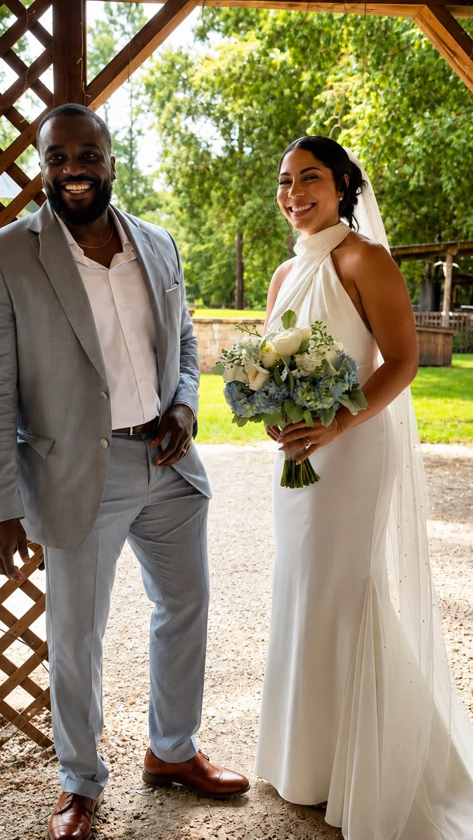 A happy bride holding a bouquet of white and light blue flowers, standing next to a groom in a light gray suit, outdoors under a wooden arbor with lush green trees in the background.