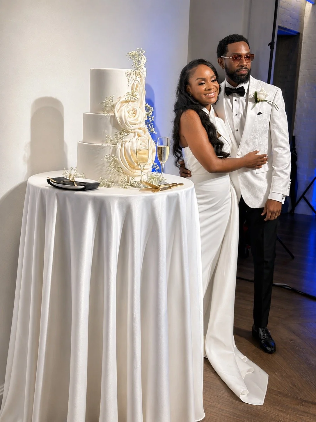 A newlywed couple standing next to a tall wedding cake on a round table, smiling and embracing, at their wedding reception.