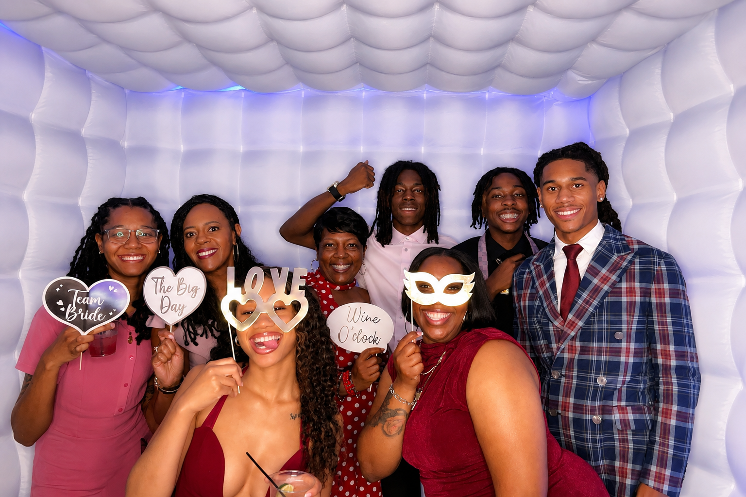 Group of people celebrating at a wedding reception photo booth, wearing party props and holding signs with messages like 'Team Bride,' 'The Big Day,' 'LOVE,' and 'Wine O'clock.'