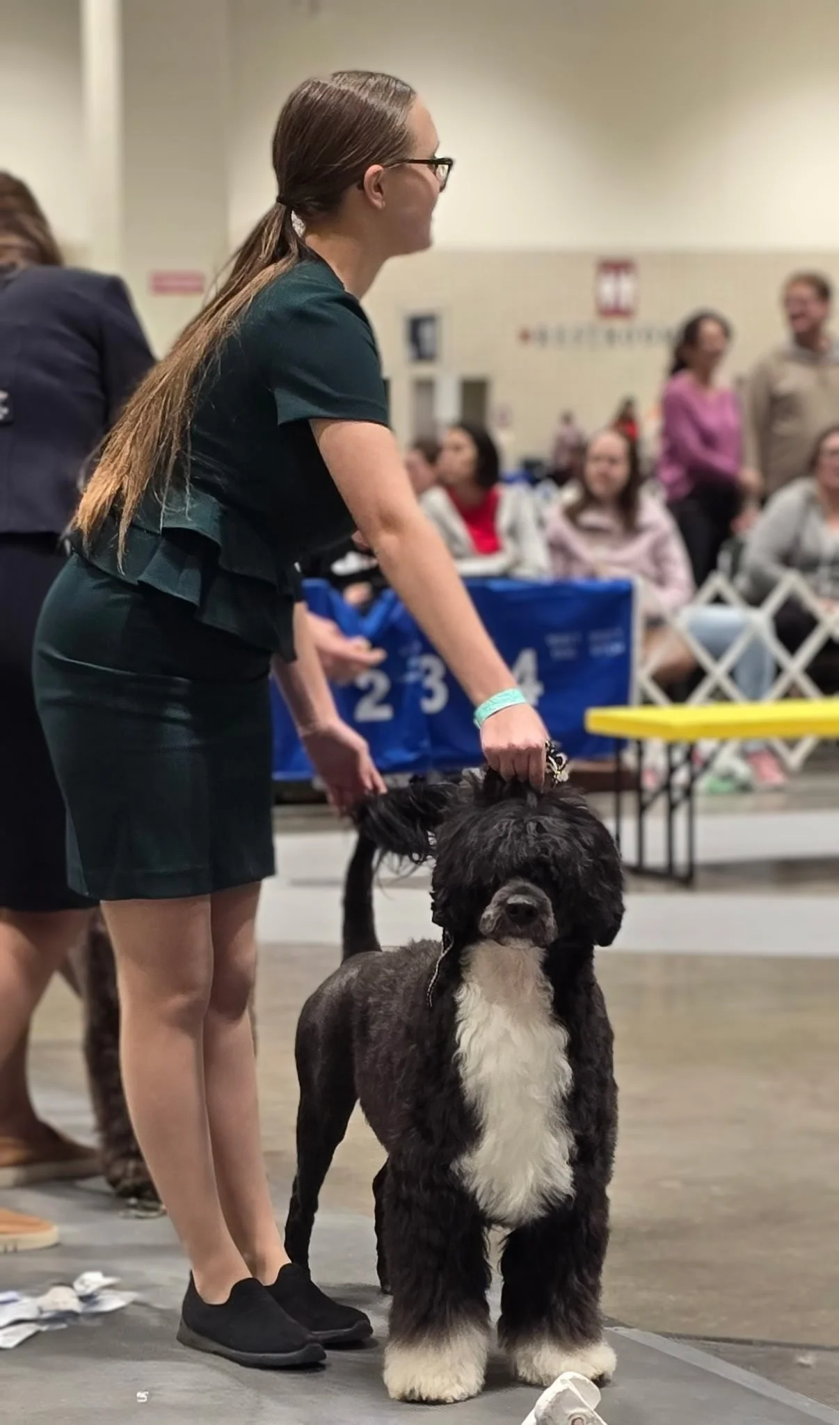 A woman with long brown hair, black glasses, wearing a black dress, holding a large black-and-white dog at an indoor dog show or competition, with several people and dog-related items in the background.