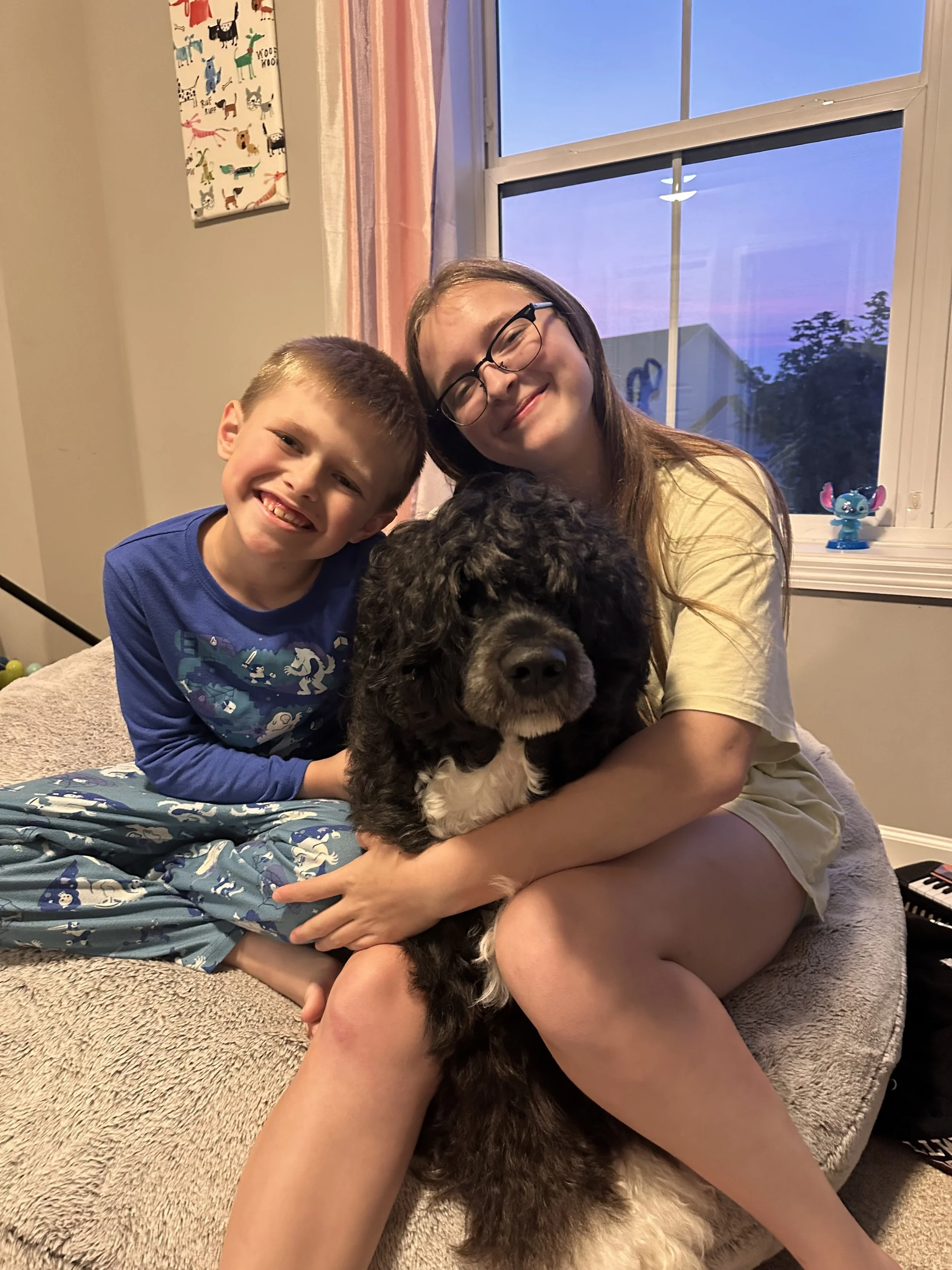 A woman with long hair and glasses, a young boy with short hair, and a large black and white dog sitting on a beige carpet in a room with a window, a pink and white striped curtain, and a colorful dog-patterned canvas on the wall.