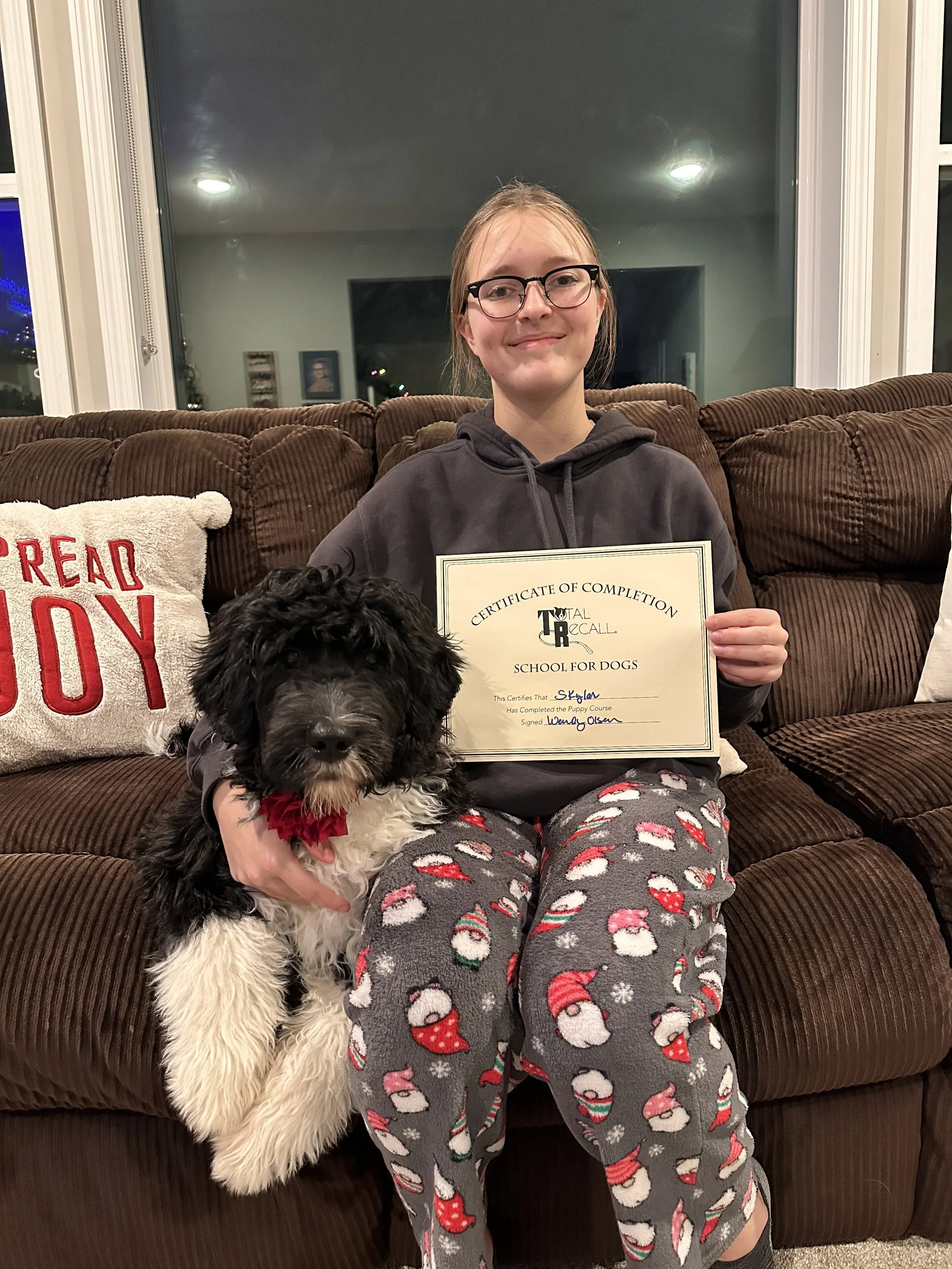A young girl with glasses holding a certificate sitting on a brown couch with a black and white puppy dog on her lap. She is wearing Christmas-themed pajamas.