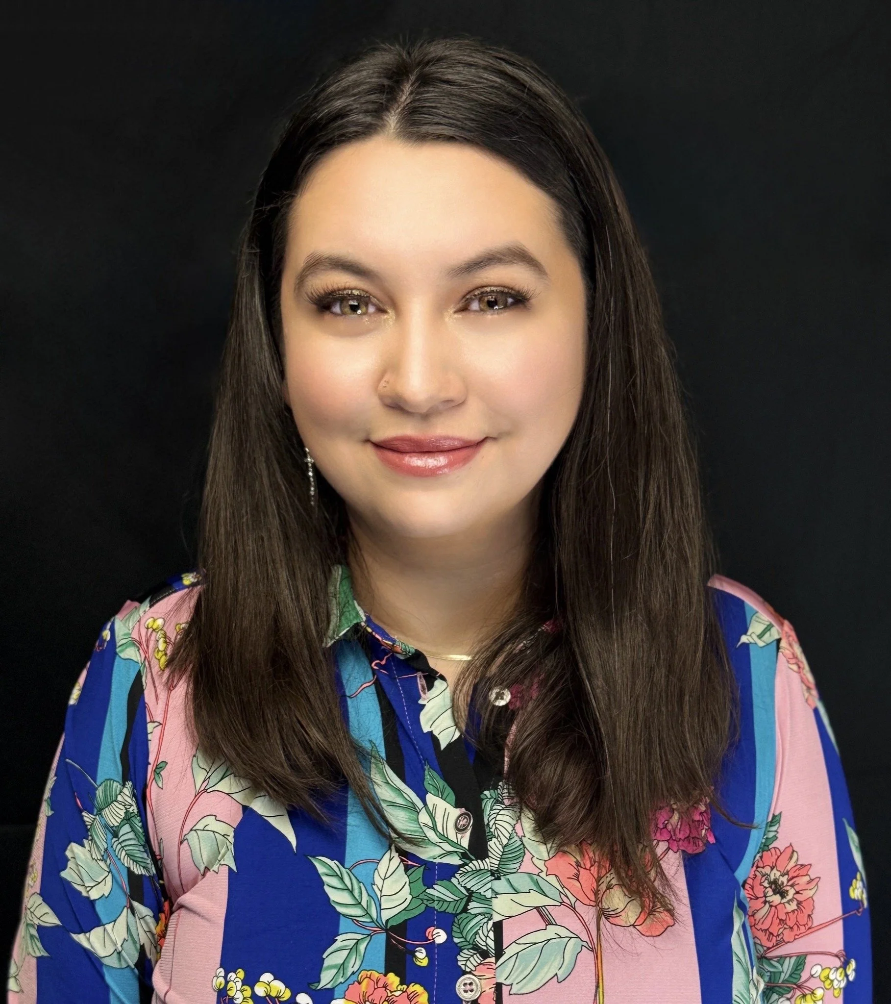 A young woman with long dark hair smiling outdoors in front of a black fence and green trees.