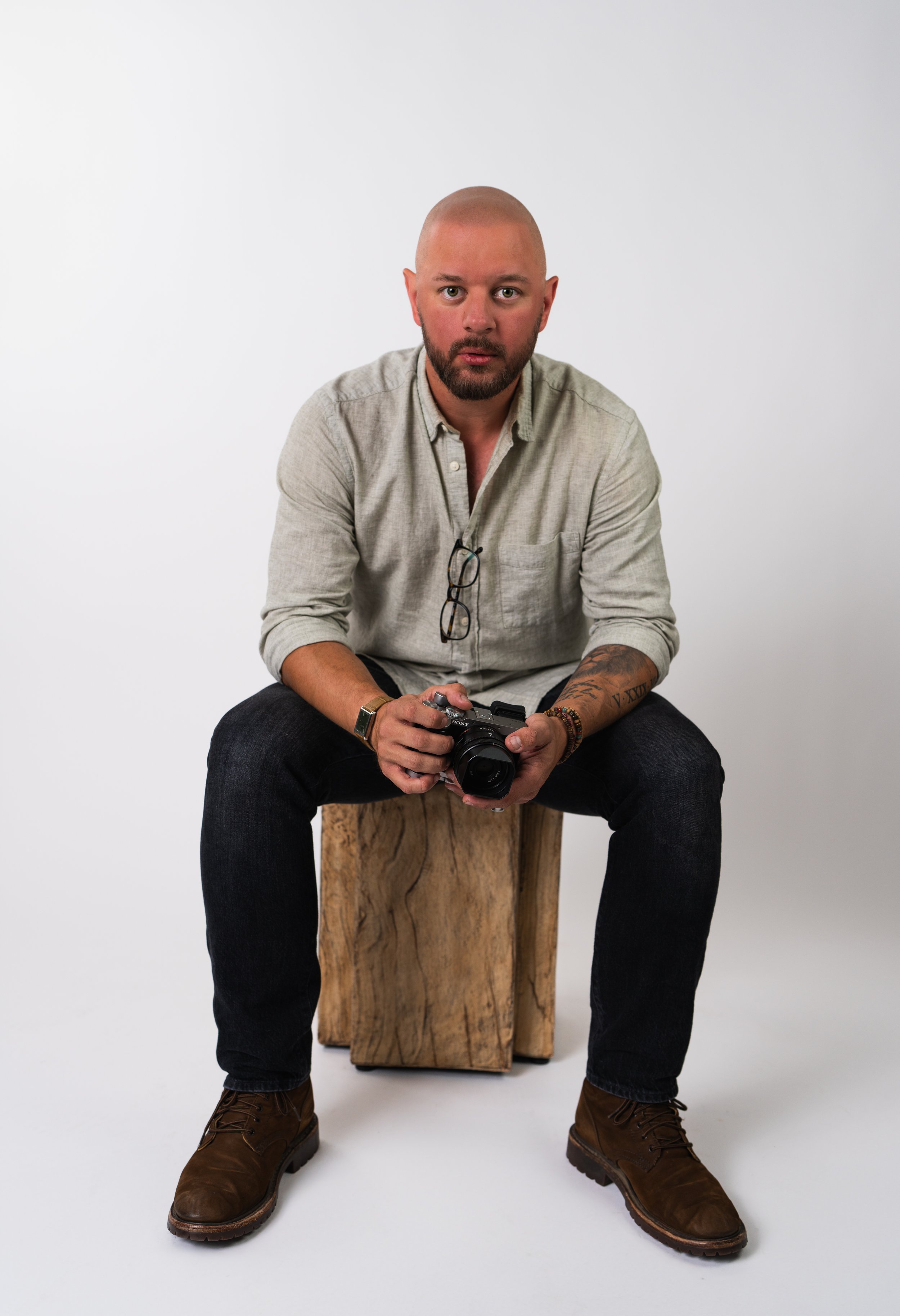 Man sitting on a wooden stool, holding a camera, wearing a light gray shirt and dark jeans, with brown shoes.