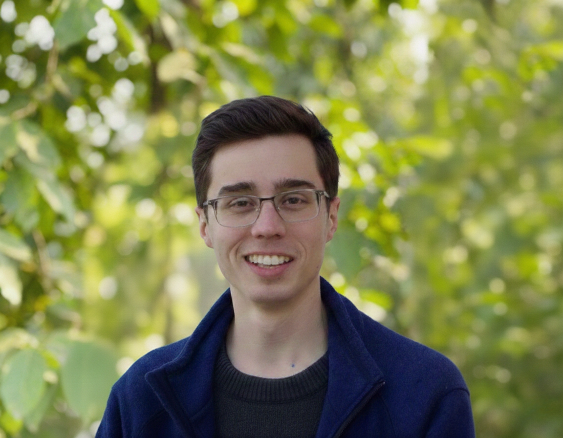A young man with glasses and dark hair smiling outdoors with trees and greenery in the background.