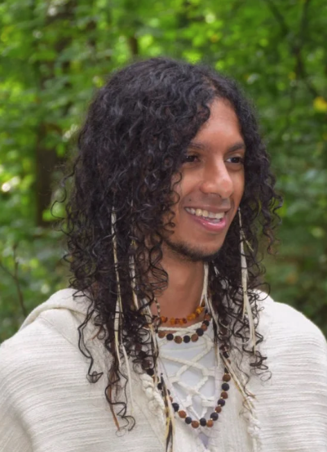 A person with long curly hair wears a white robe and beaded necklaces, smiling outdoors with green foliage background.