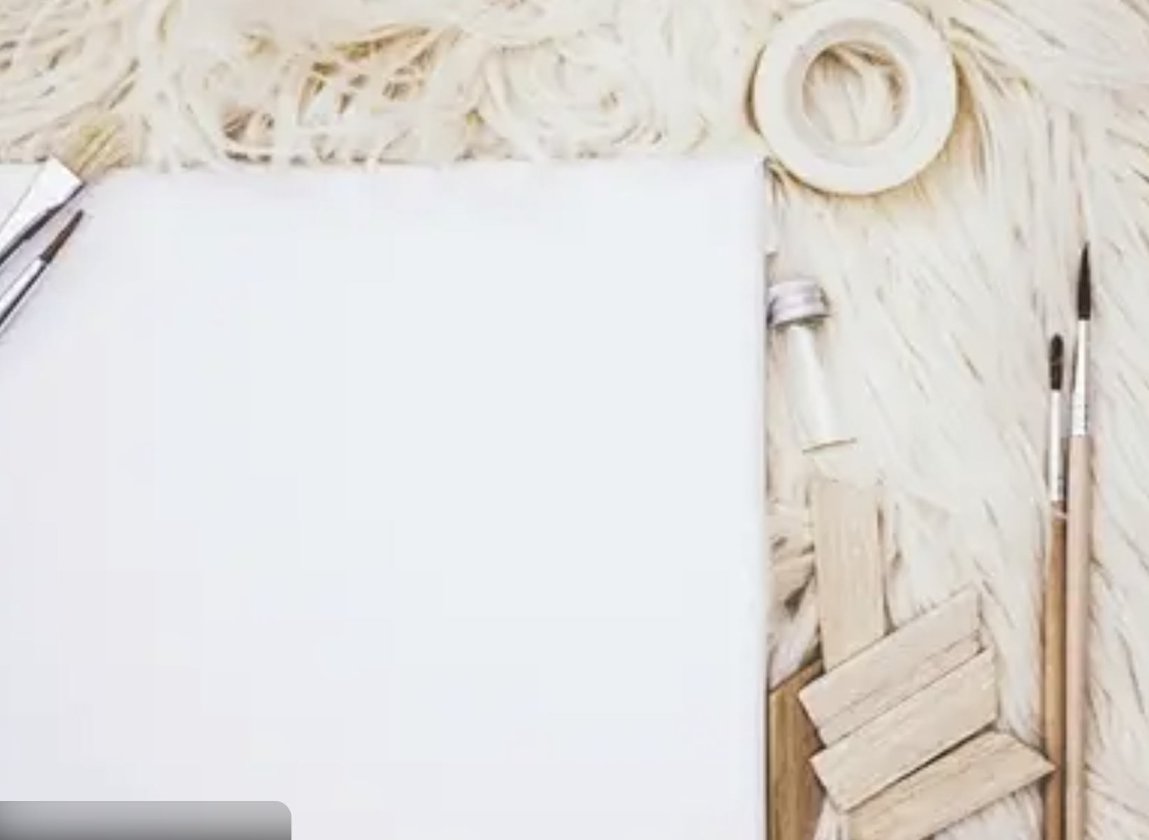 A blank white canvas on a light wooden table with art supplies around it, including paintbrushes, painter's tape, and a small wooden palette.