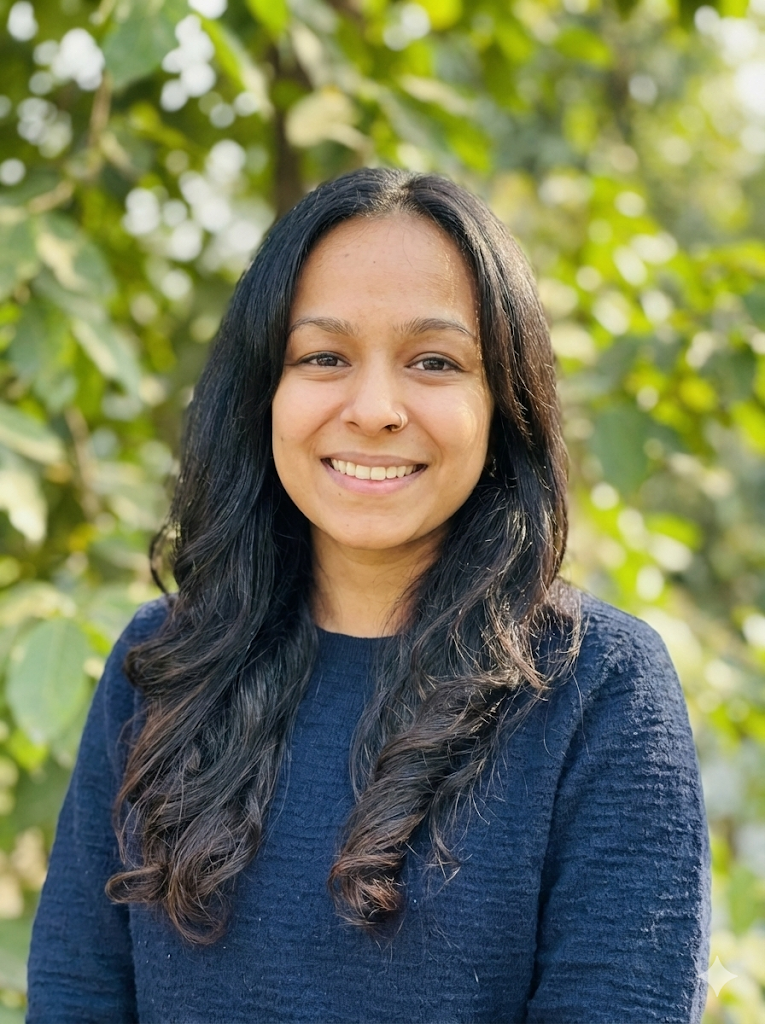 A woman with long black hair, wearing a navy blue sweater, smiling outdoors with greenery in the background.