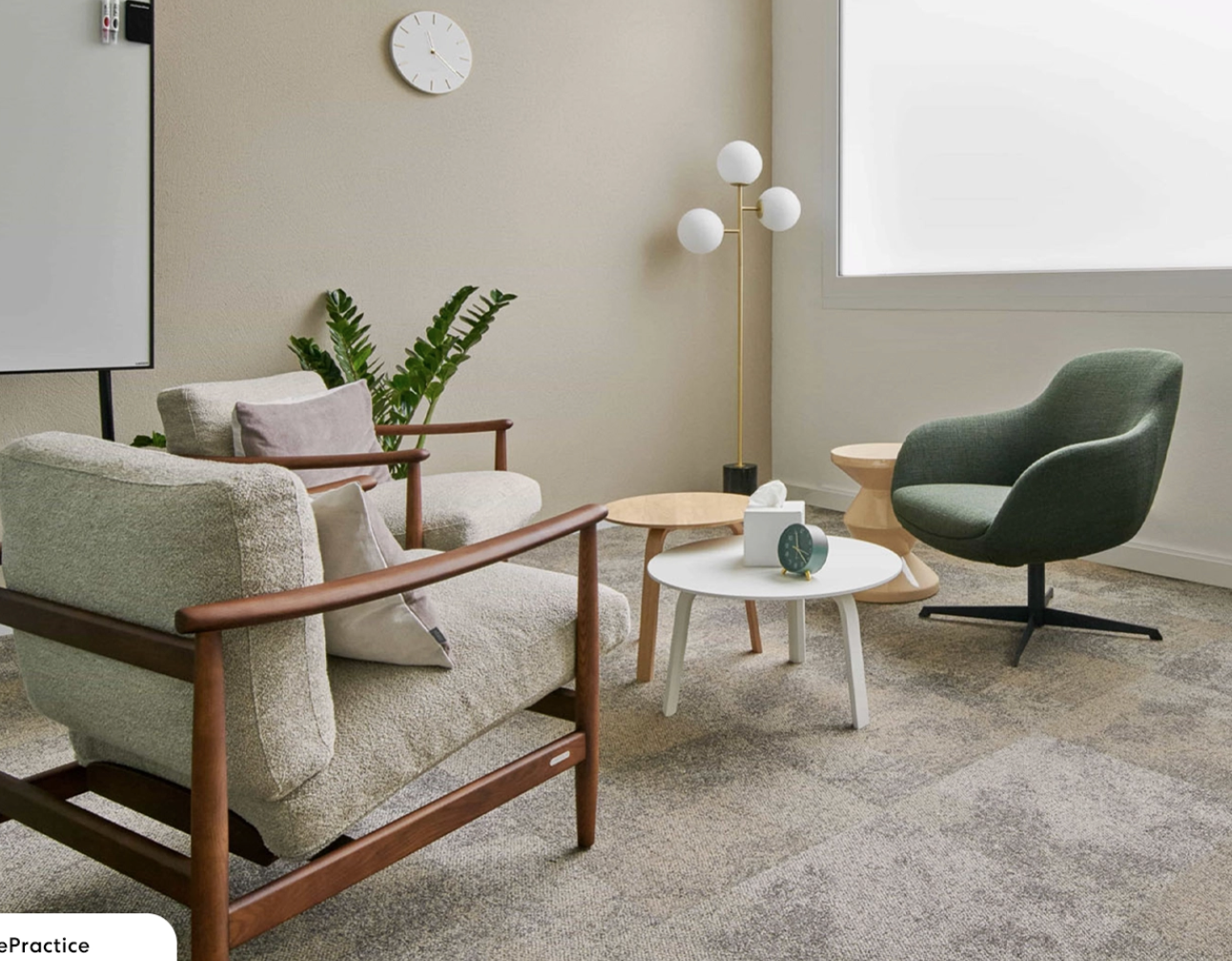 A modern office or waiting room with a beige sofa, dark green armchair, three nested round tables, a potted plant, a floor lamp with white globes, a wall clock, and a large window with frosted glass.