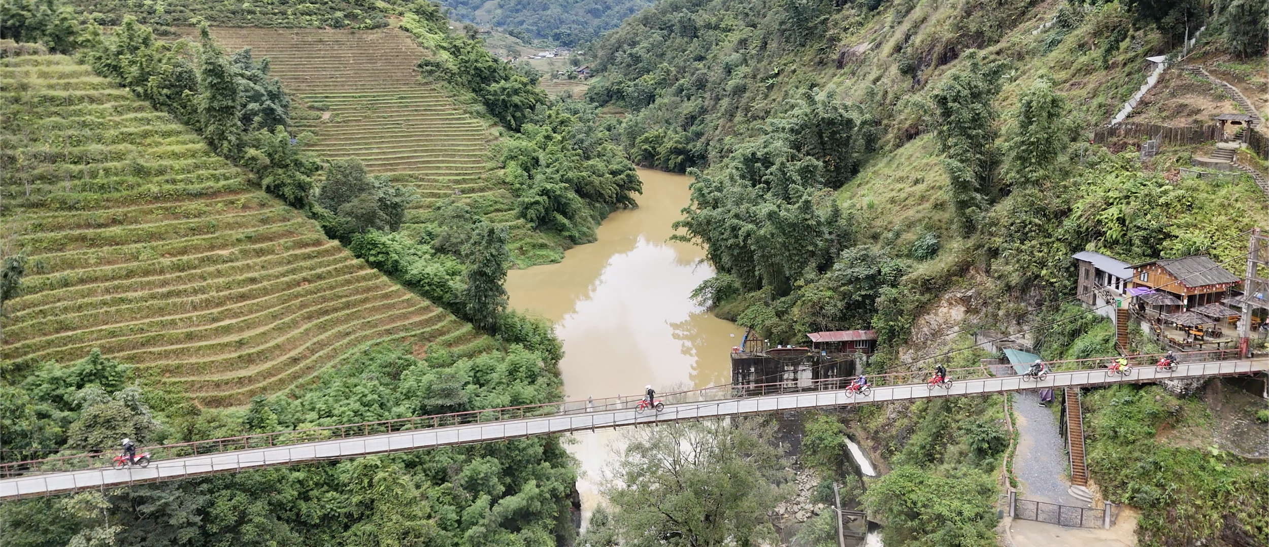 Motorcycle riders crossing a narrow bridge over a river surrounded by jungle and terraced rice fields on a Vietnam adventure motorcycle tour