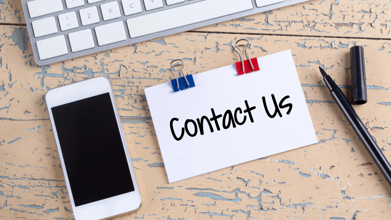 Photo of a workspace with a computer keyboard, a smartphone with a black screen, a pen with an open cap, and a white note card with the words "Contact Us" attached to a rough wooden surface with two clips, one blue and one red.