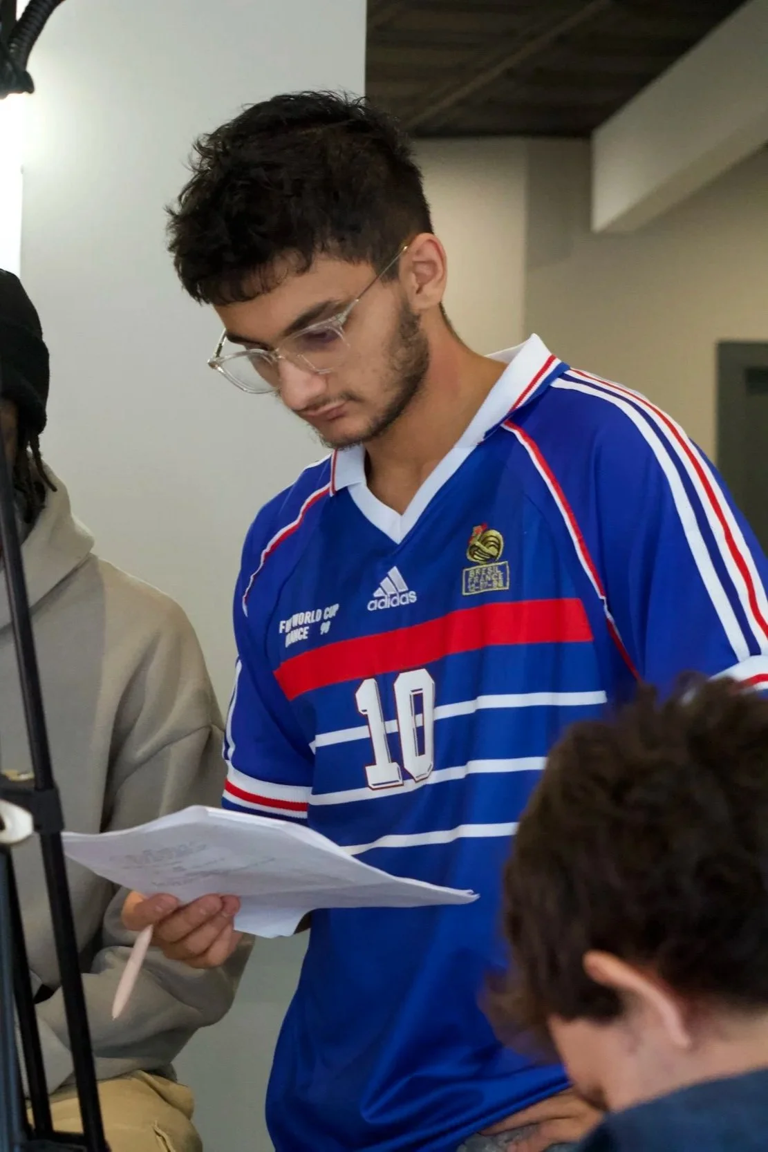 A young man wearing glasses and a blue sports jersey with the number 10 is reading a sheet of paper while standing.