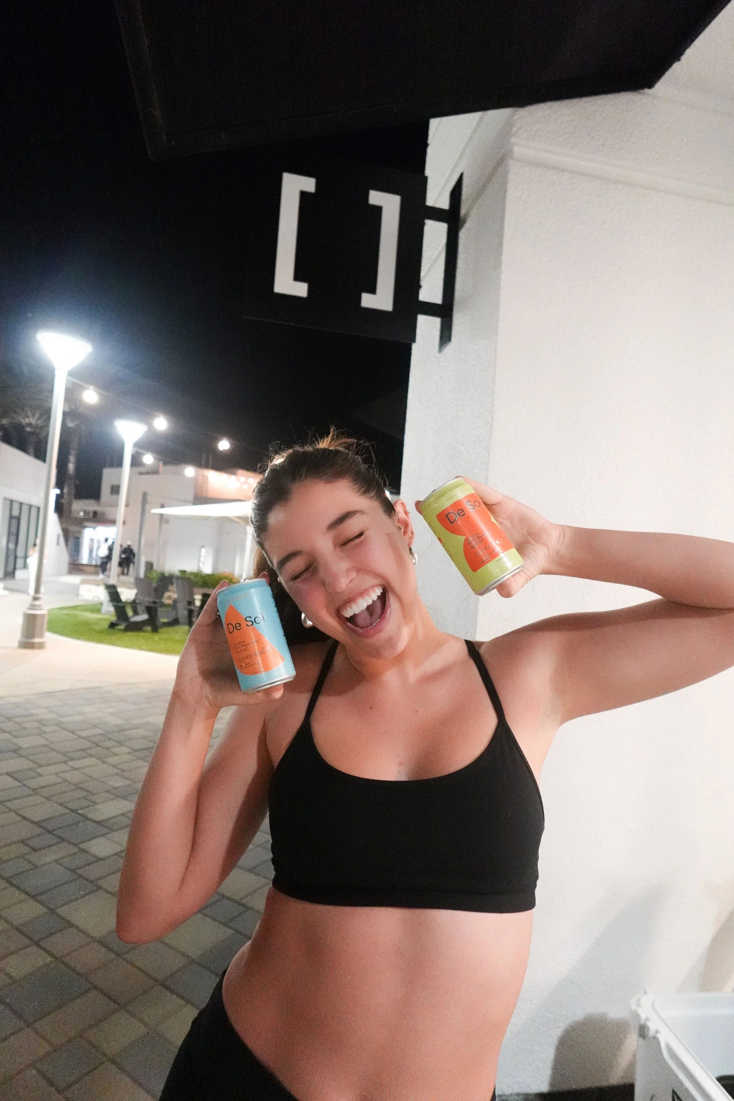 A young woman in a black sports bra holding two cans of De Soi beverage, smiling and posing outdoors at night.